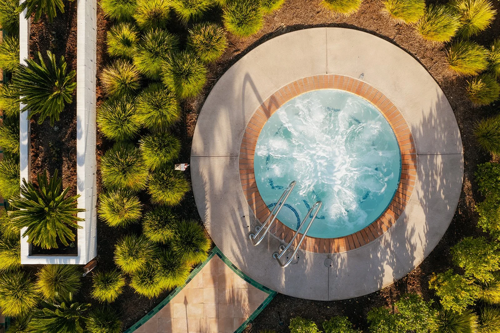 An aerial view of a hot tub with bubbling water, surrounded by concrete and brick, next to a landscaped area with green plants and trees.