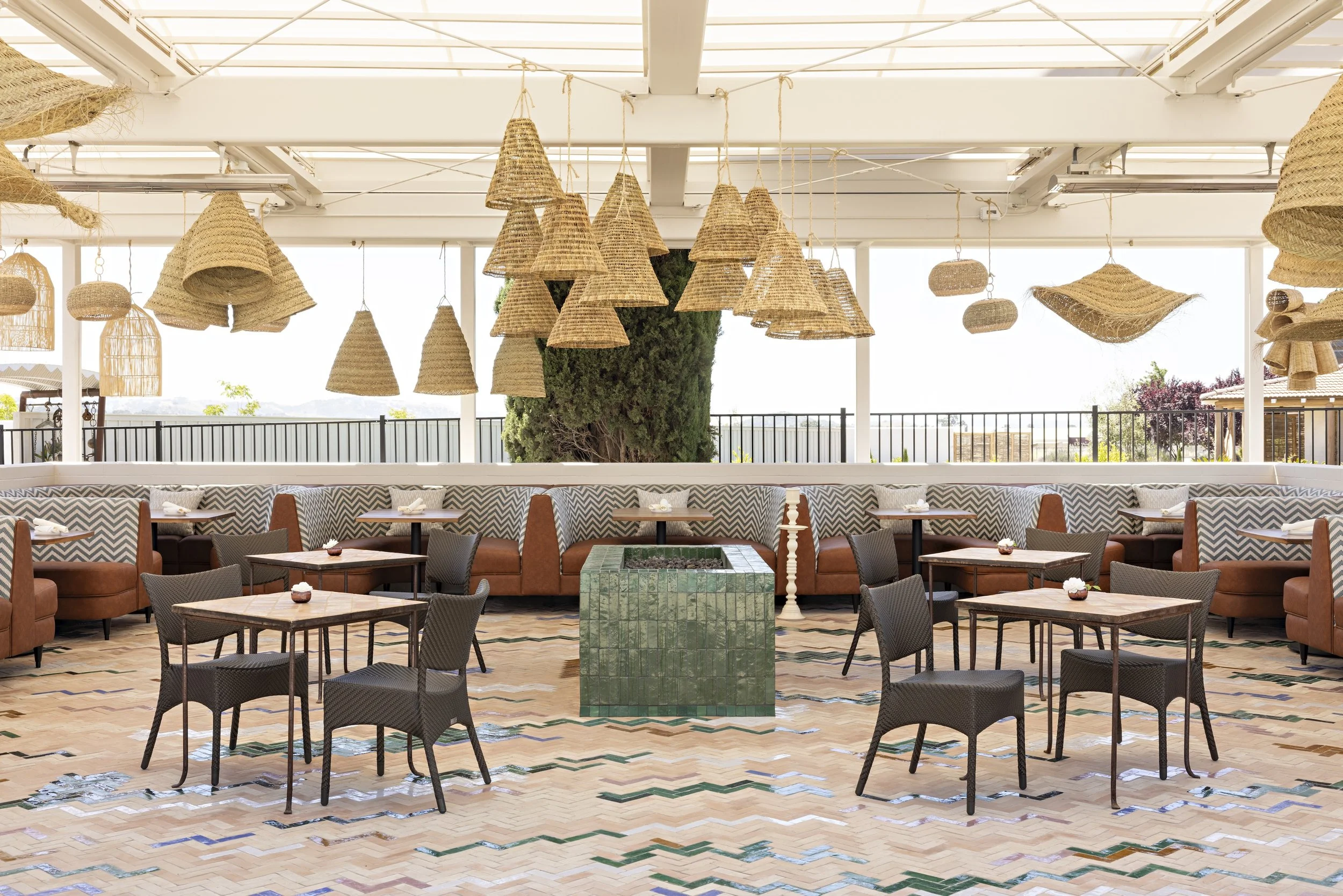 Indoor dining area with numerous tables and chairs, pendant woven straw light fixtures hanging from the ceiling, and a large plant in the center. The space has large windows and a railing outside.
