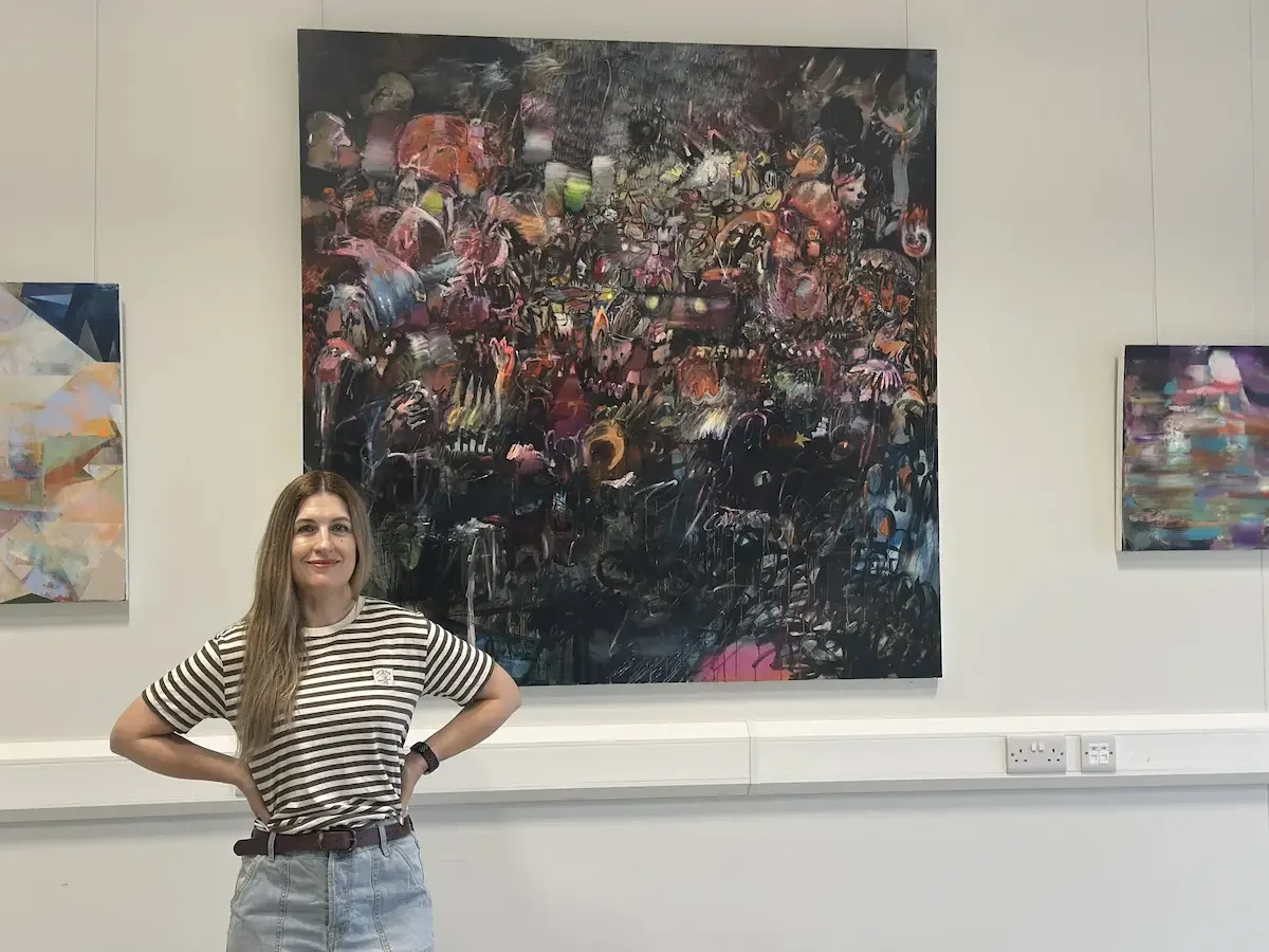 A women in a stripy top sat in front of a massive painting in an art gallery.