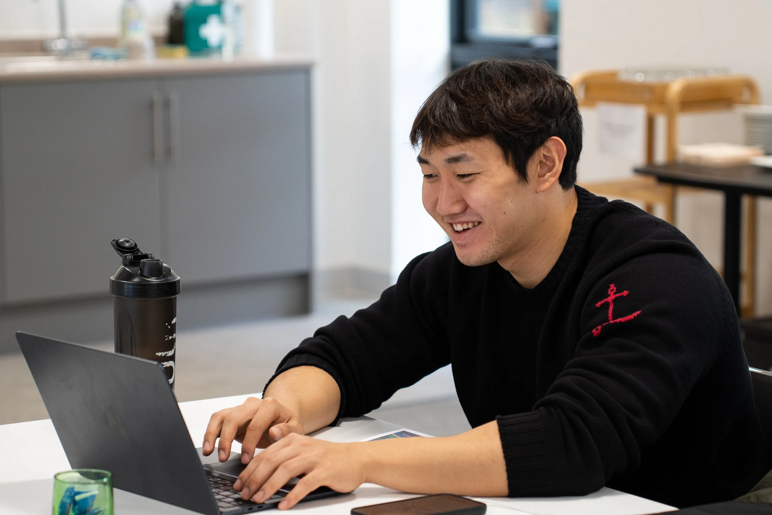 A smiling man in a black sweater sitting at a white desk, typing on a laptop. He appears engaged and happy while working in a bright, modern office kitchen or breakroom area. A water bottle and phone are on the desk beside him.