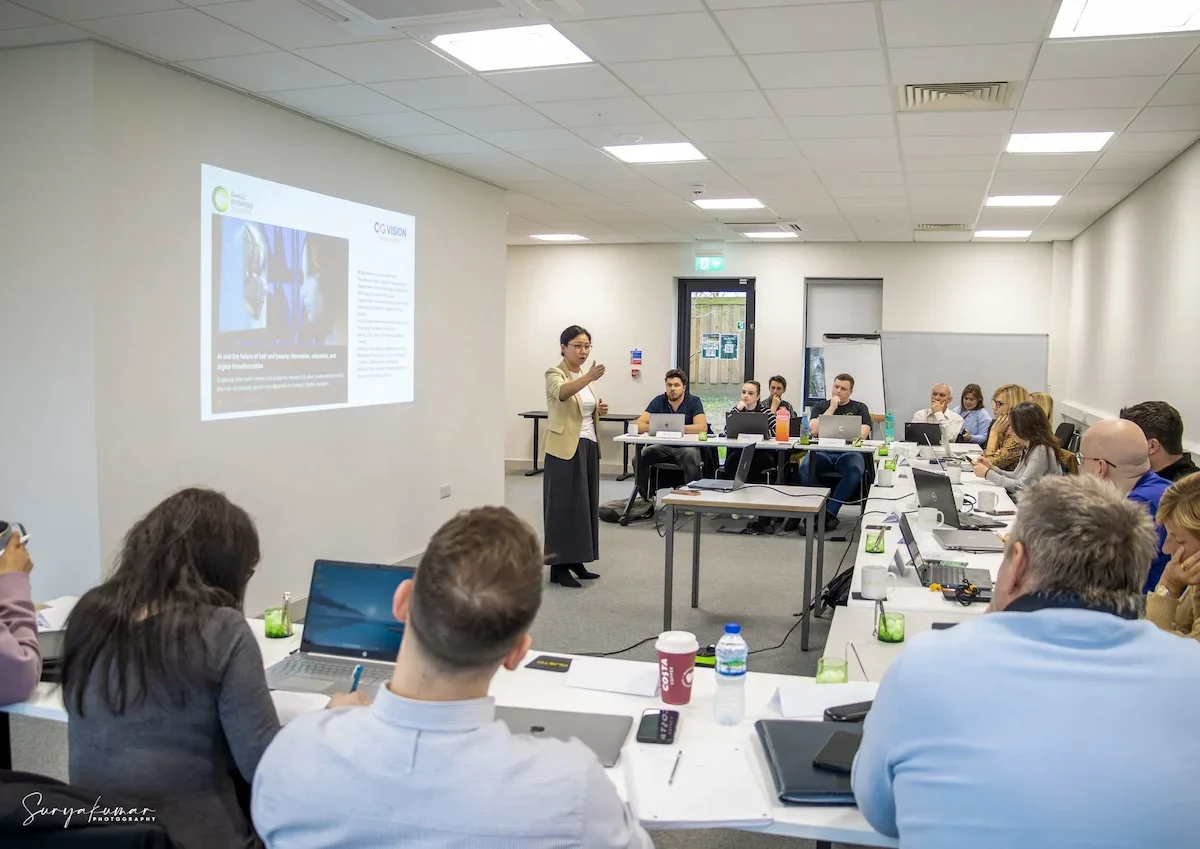 A group of approx. 16 people sat in a training room, u shaped layout, with laptops, notebooks and pens