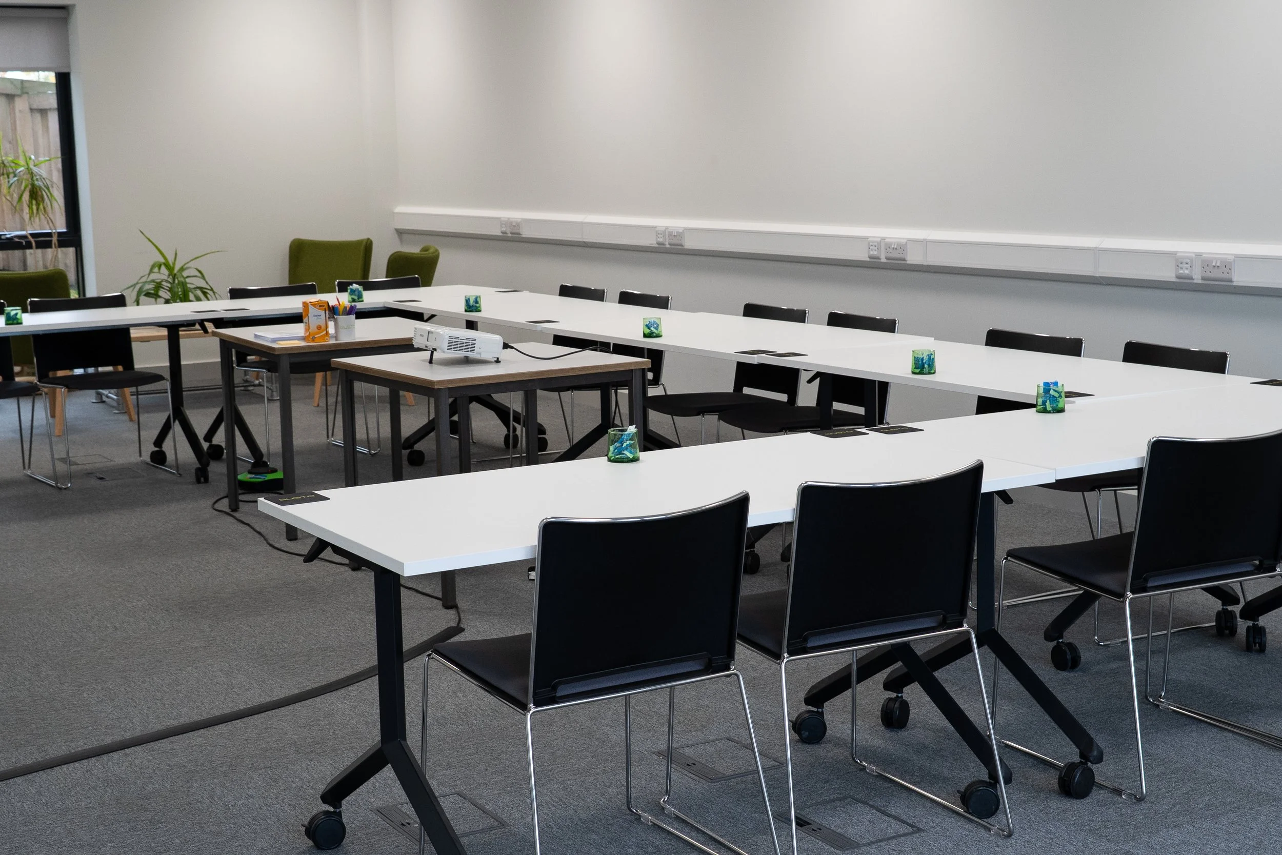 An empty, professional training room hire near Newark, featuring white desks in a U-shape configuration, black chairs, and natural lighting.