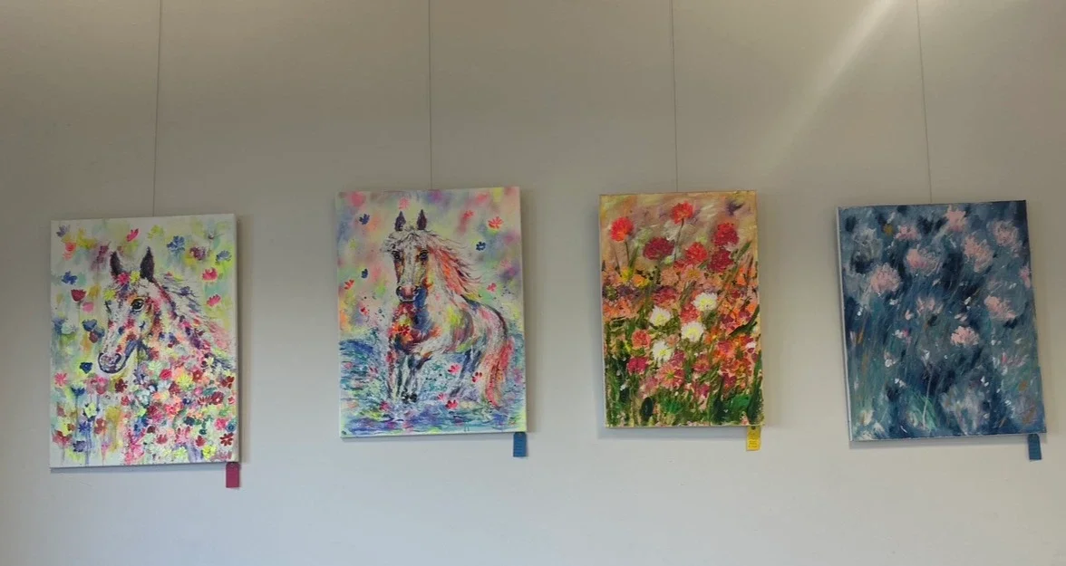 4 painted pictures hanging on a white wall, all pictures contain really bright and vibrant colours. Two paintings are of a horse and the other two are of flowers.
