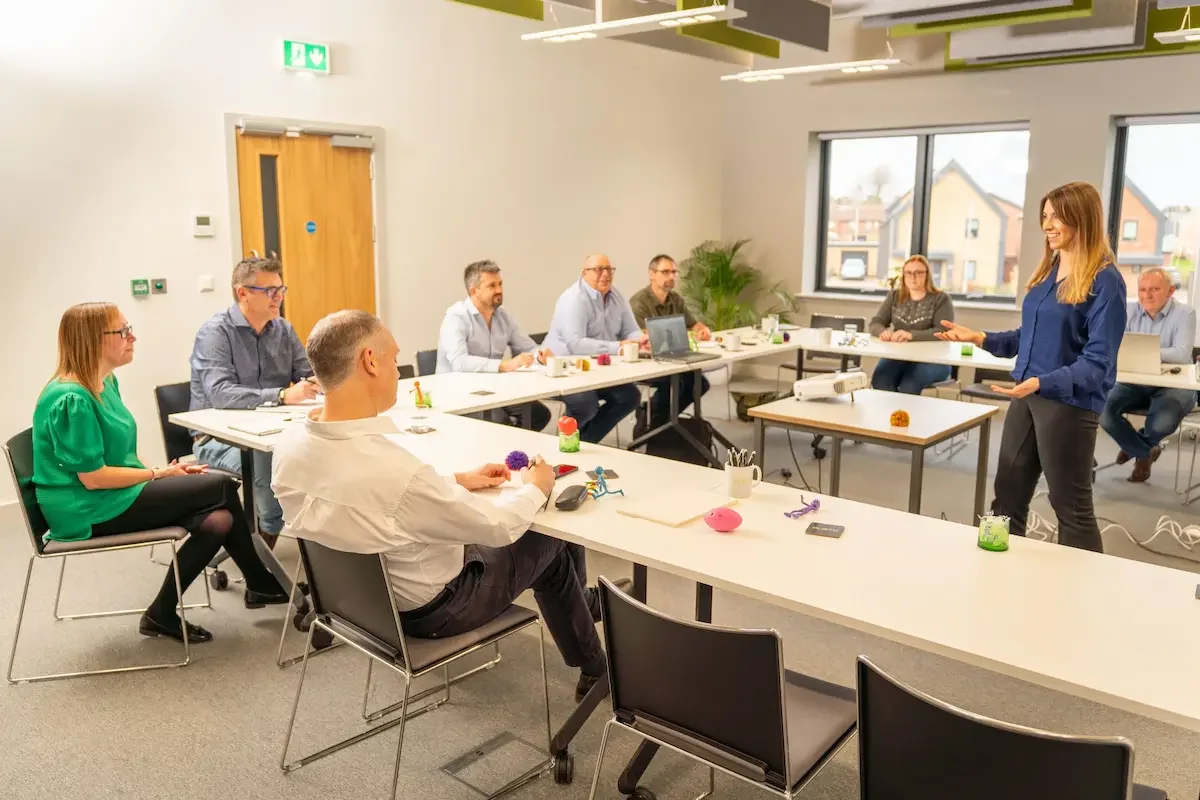 A group of 8 people sat in a training room, u shape layout with another person delivering a presentation. Everyone is smartly dressed in the room, which has large windows.