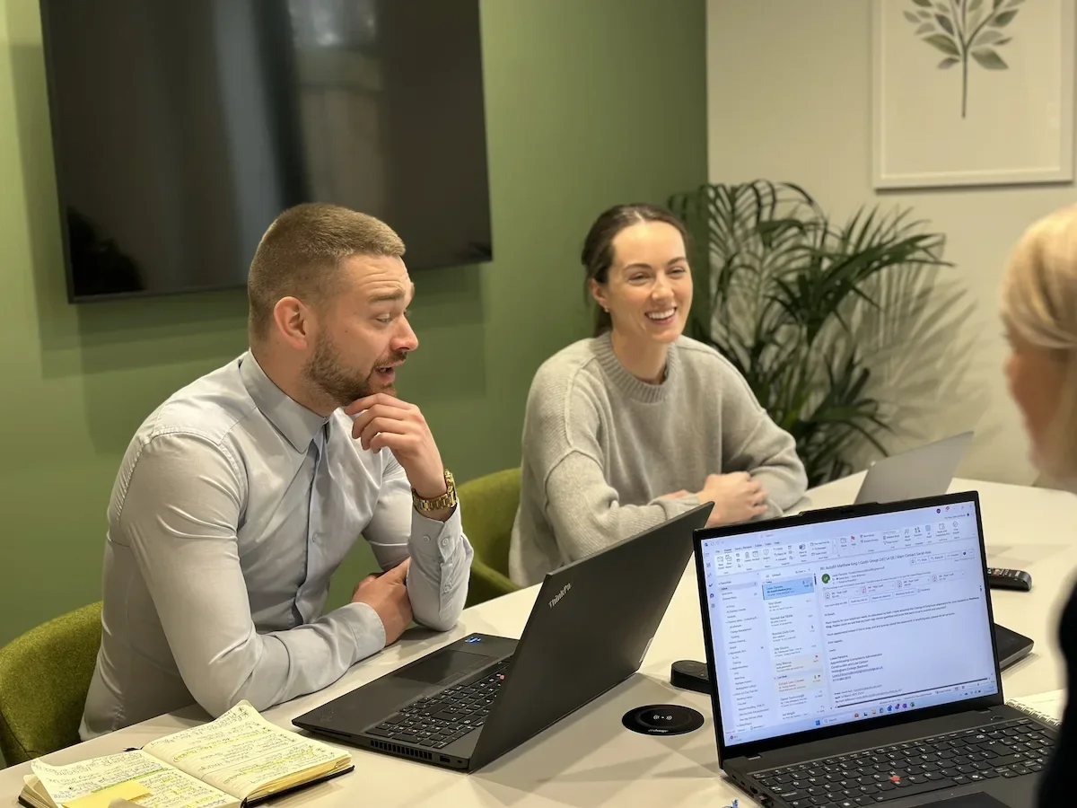 Two people laughing and smiling while sat in the Gusto House Meeting Room, with a green wall and a smart TV behind them