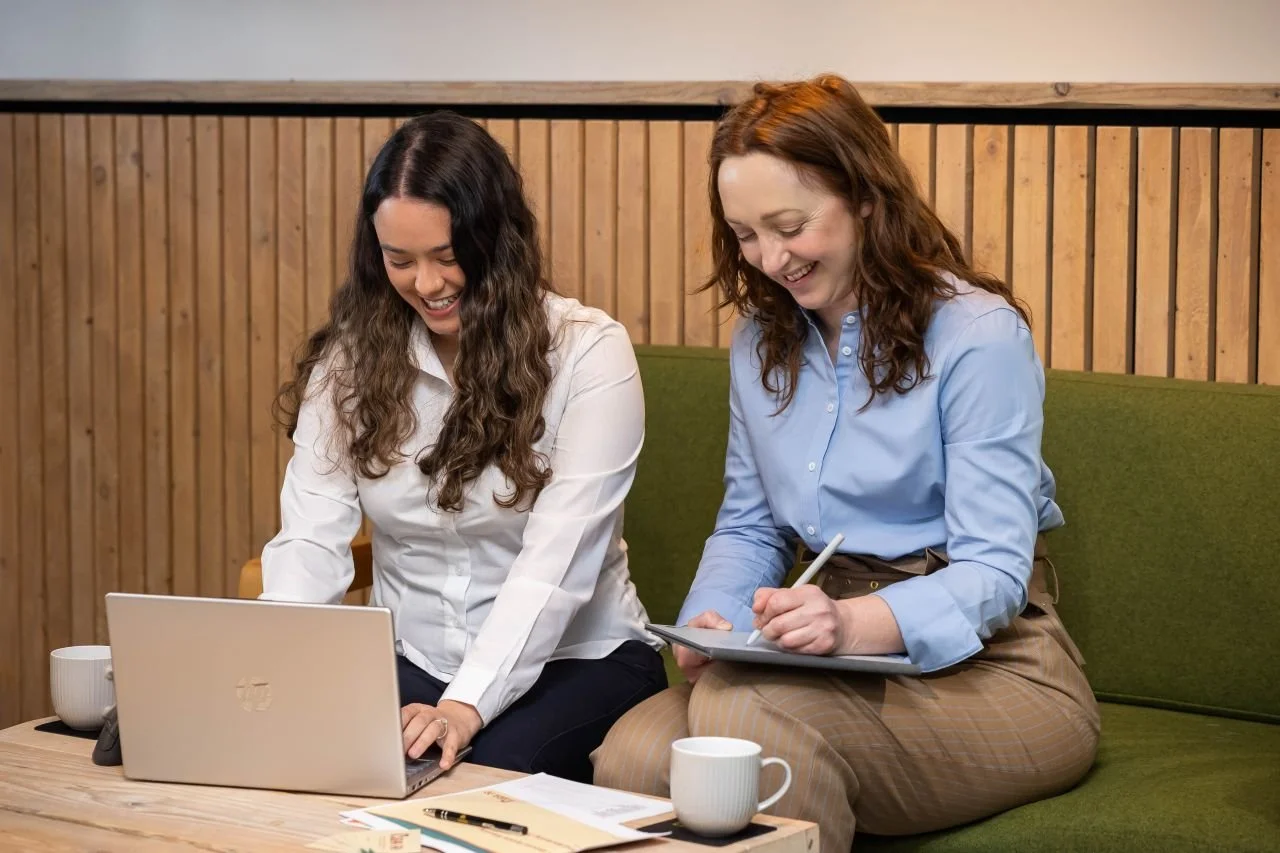 Two coworkers laughing and smiling at the coworking space at Gusto House Collingham. They are sat on a green work bench with wooden panelling behind them. One has a MacBook and t he other is writing on an iPad