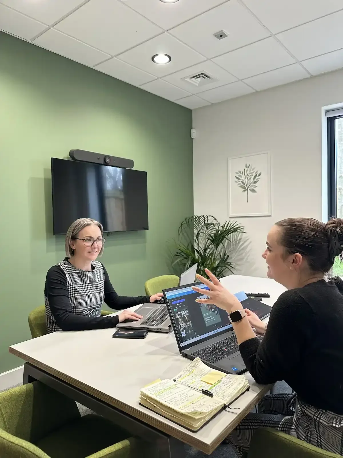 Two women in a meeting room with laptops, notebooks, and a TV screen on the wall, engaged in discussion.