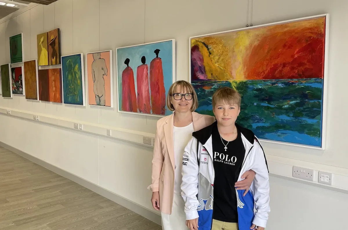 A women with short blonde hair and glasses stood with her arm around a boy with blonde hair and blue eyes. They are stood in front of an art gallery filled with big, vibrant and colourful artwork