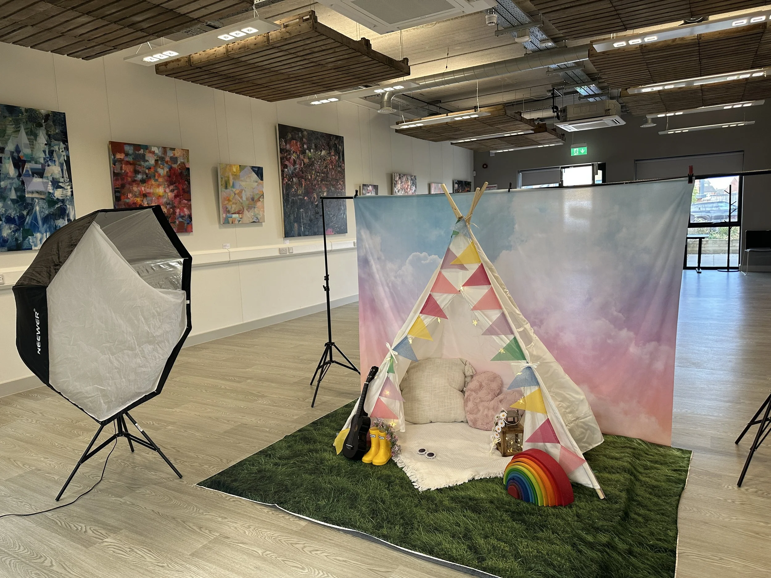 A professional photography setup in a large gallery room featuring a softbox light, a "grass" floor mat, and a white teepee decorated with colorful bunting against a sky-blue backdrop.