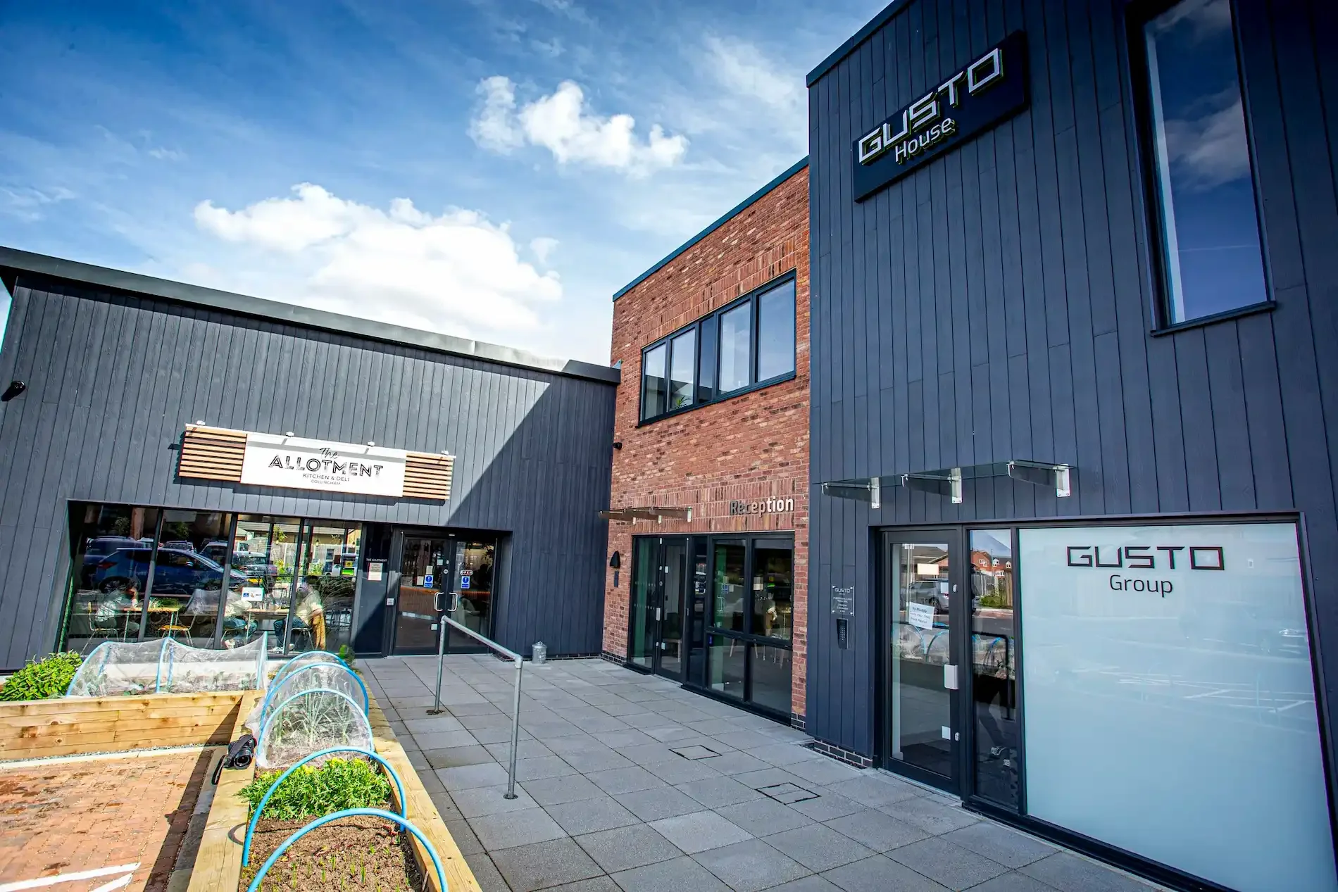 Outside of the main entrance to a modern office called ' GUSTO House'. This sits right next door to a cafe and deli called The Allotment