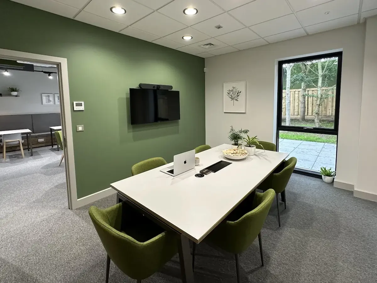 A modern meeting room which contains a large window, a smart tv on a green wall, and table surrounded by 6 green chairs