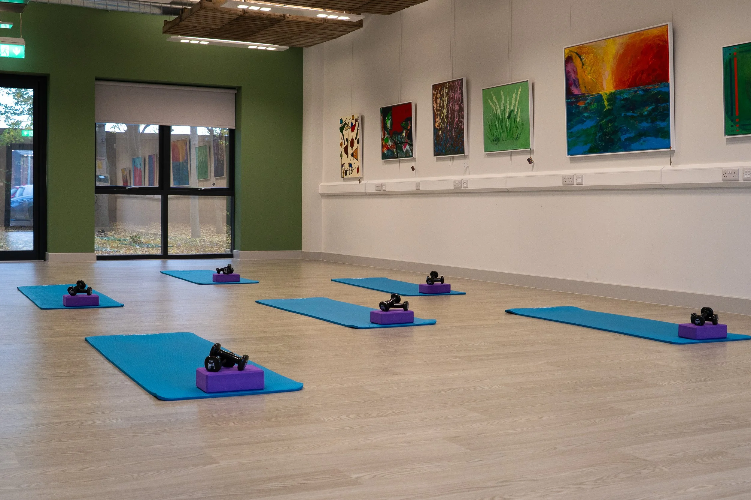 An art gallery with artwork displayed on the walls, a wooden ceiling with lighting fixtures, large glass doors allowing natural light in, and hardwood flooring.