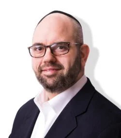 Portrait of a middle-aged man with glasses, a beard, wearing a dark suit and white shirt, against a plain background.