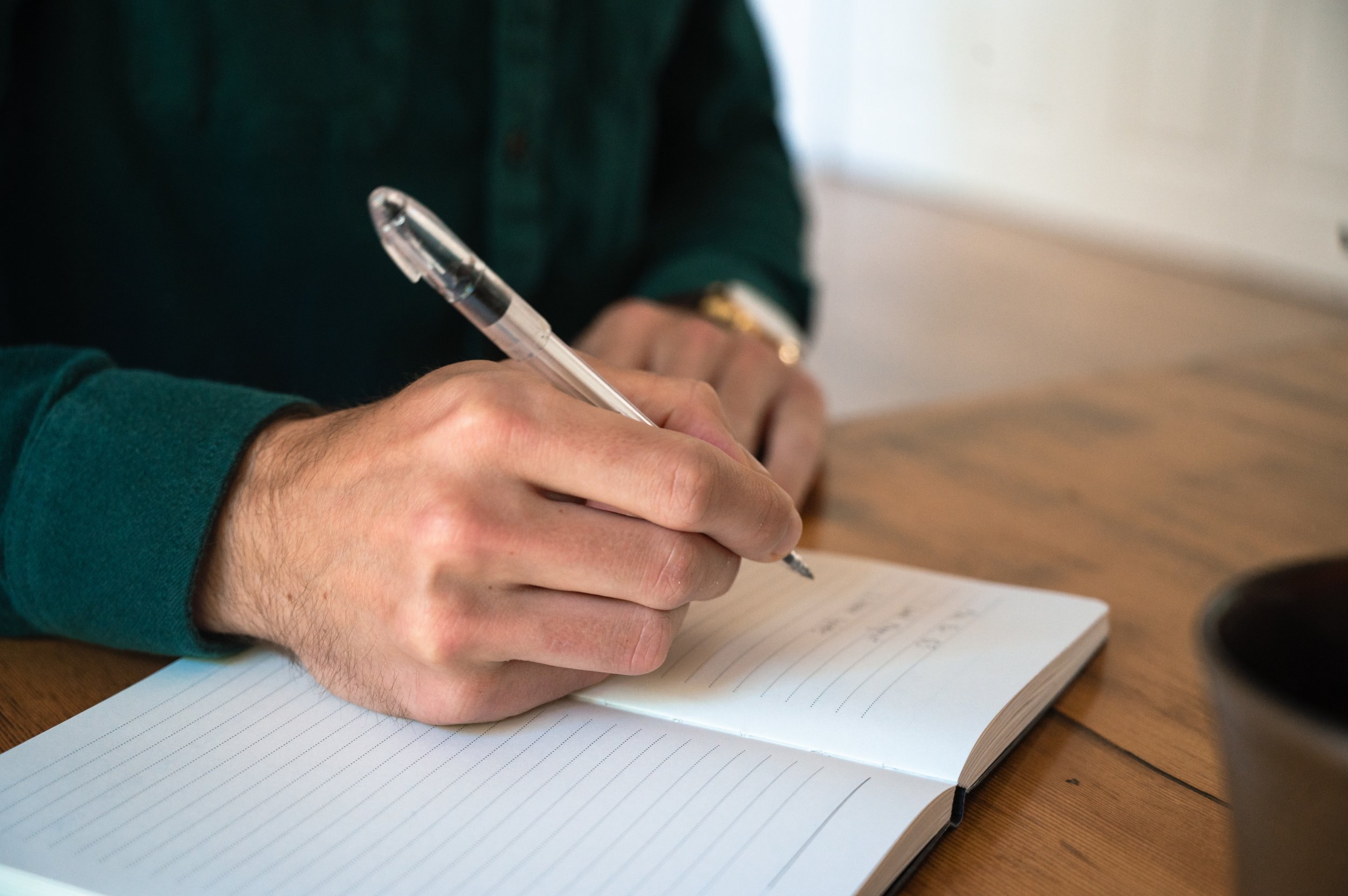 A person in a green sweater is writing in a notebook with a clear ballpoint pen.