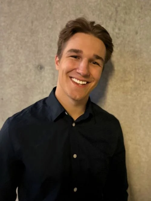A young man with short brown hair wearing a dark button-up shirt, smiling, standing against a textured beige wall.