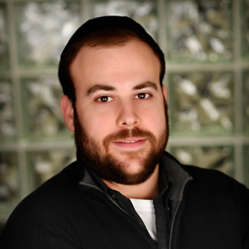 A man with a beard and short dark hair, wearing a black jacket, in front of a window with trees outside.