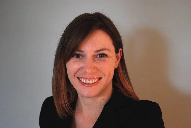 A woman with shoulder-length brown hair smiling at the camera, wearing a black top against a plain light-colored wall.