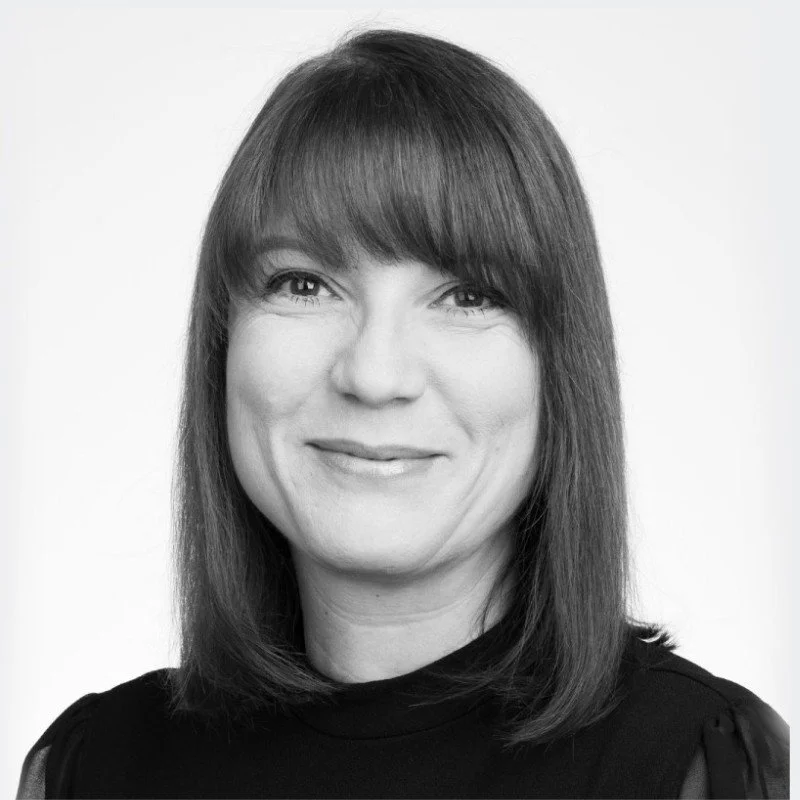 Black and white portrait of a woman with shoulder-length straight hair and bangs, smiling softly, wearing a dark top with a high neckline.