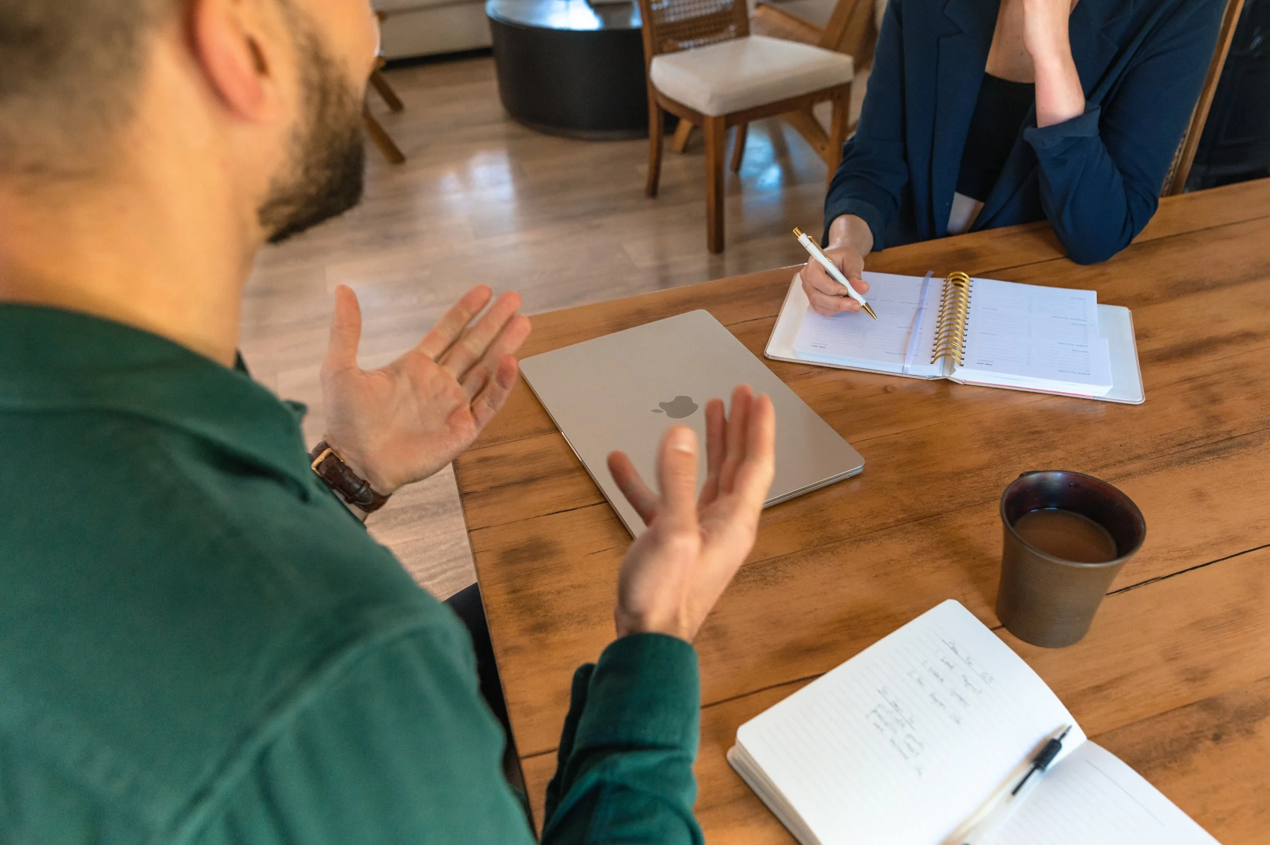 A person with a beard and glasses wearing a green sweater is talking to another person, who is holding a pen and sitting at a wooden table. The person with the pen has notebooks and a laptop on the table, with a cup of coffee nearby.
