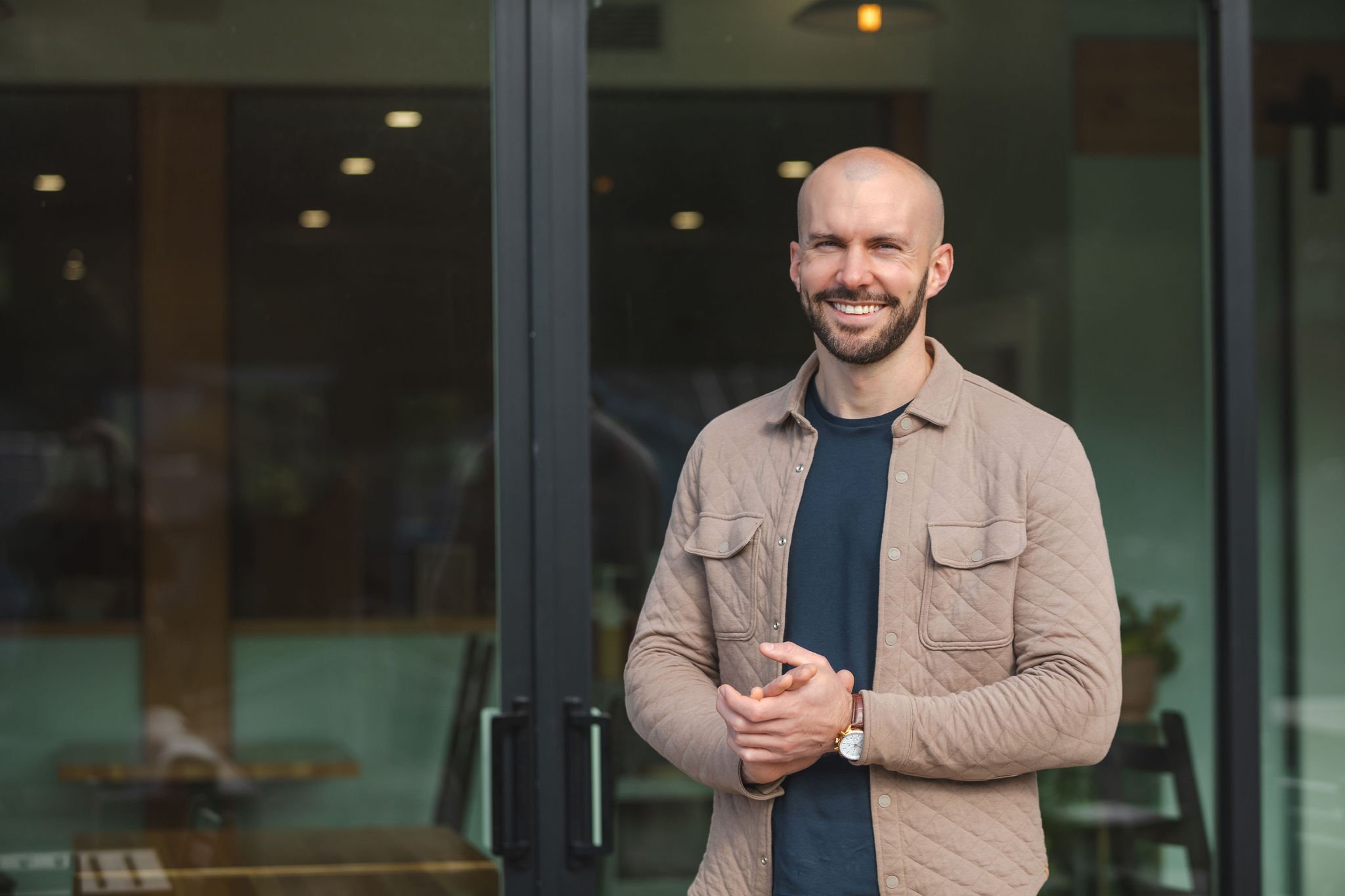 A smiling man with a beard and a watch stands outside in front of glass doors.