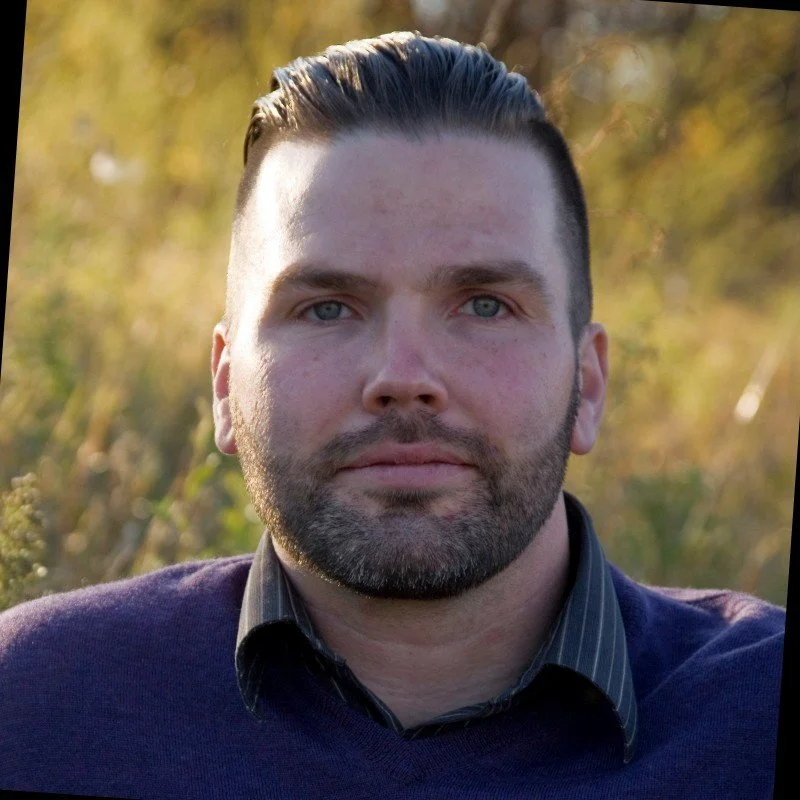 A man with slicked-back hair and a beard outdoors with blurred foliage in the background.