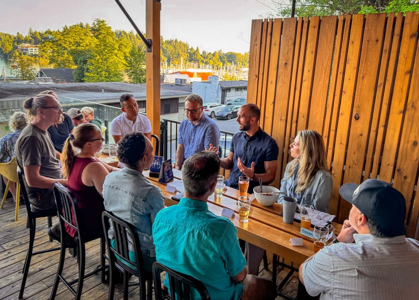 A group of people gathered around a long wooden table on an outdoor deck, engaged in a discussion, with drinks and bowls on the table. The setting is a casual outdoor venue with a wooden fence and a view of trees and parked cars in the background.