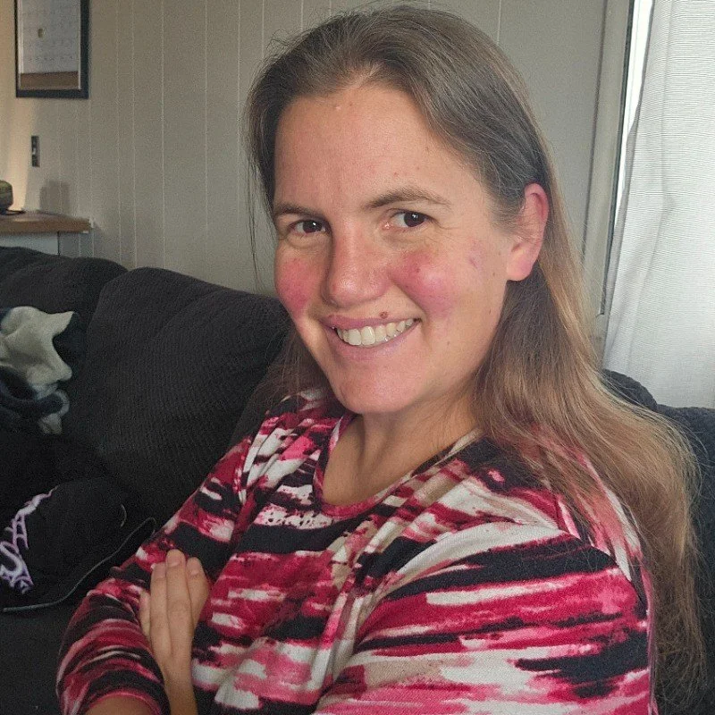 A woman with brown hair, smiling with arms crossed, sitting on a couch in a living room.