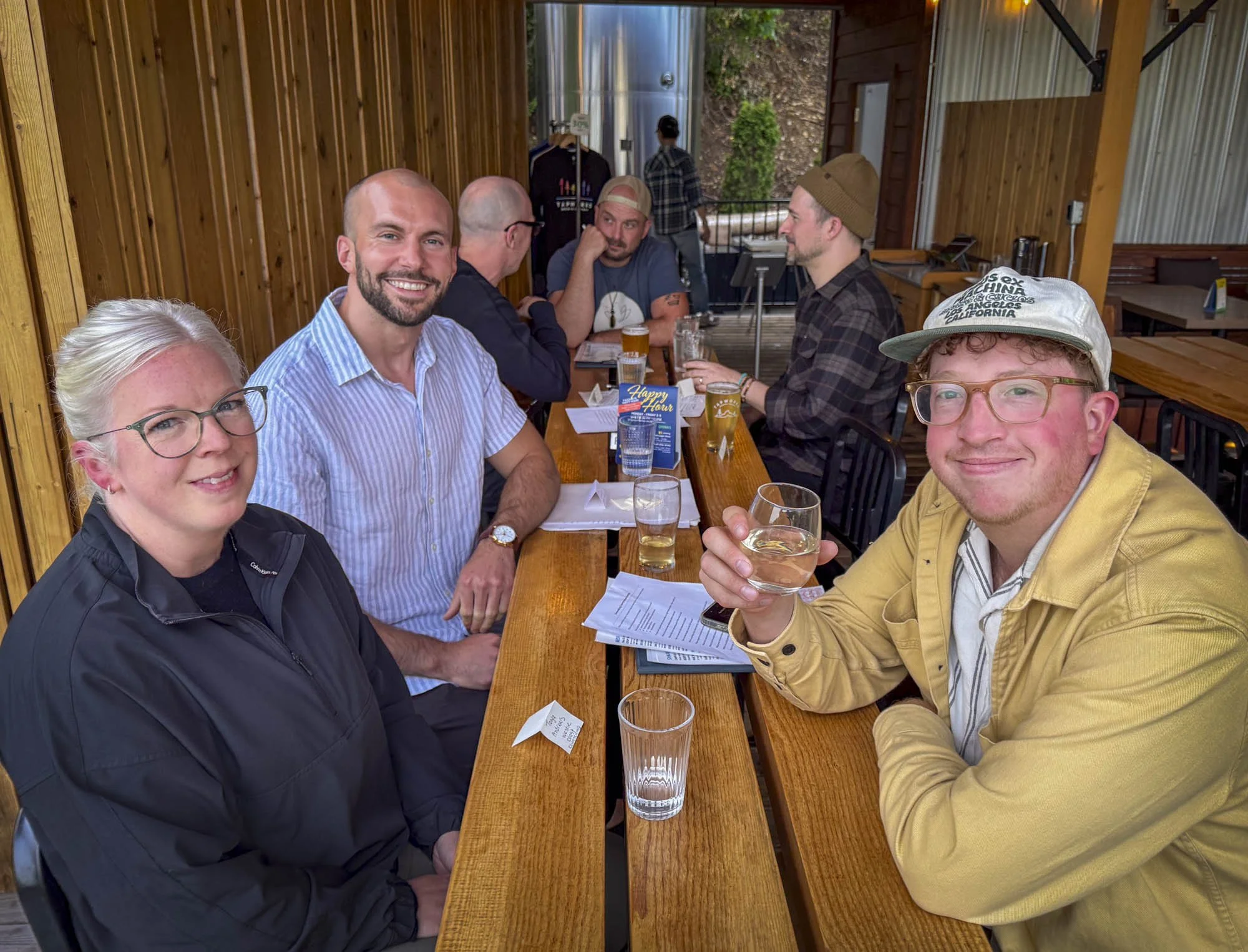 Group of six people sitting and standing at a long wooden table in a restaurant or pub, with some drinks and menus in front of them. The setting has wooden walls and a partially outdoor feel, with some green foliage visible outside. They appear to be