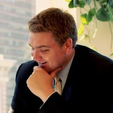 A young man in a business suit smiling thoughtfully with his hand on his chin, sitting near a window with green plants in the background.