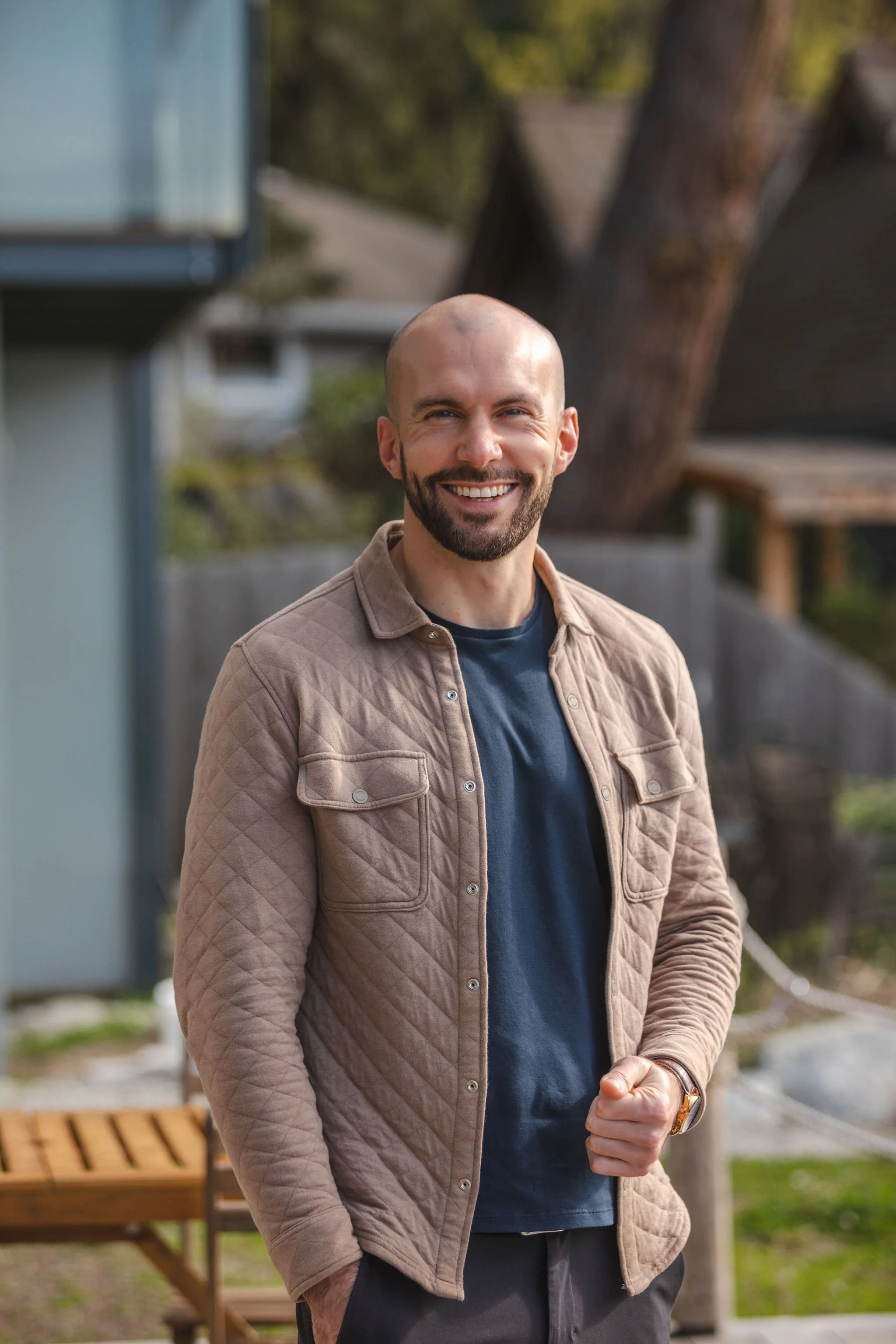 A smiling man with a beard and a shaved head standing outdoors in front of trees and a house, dressed in a beige quilted jacket over a blue shirt.