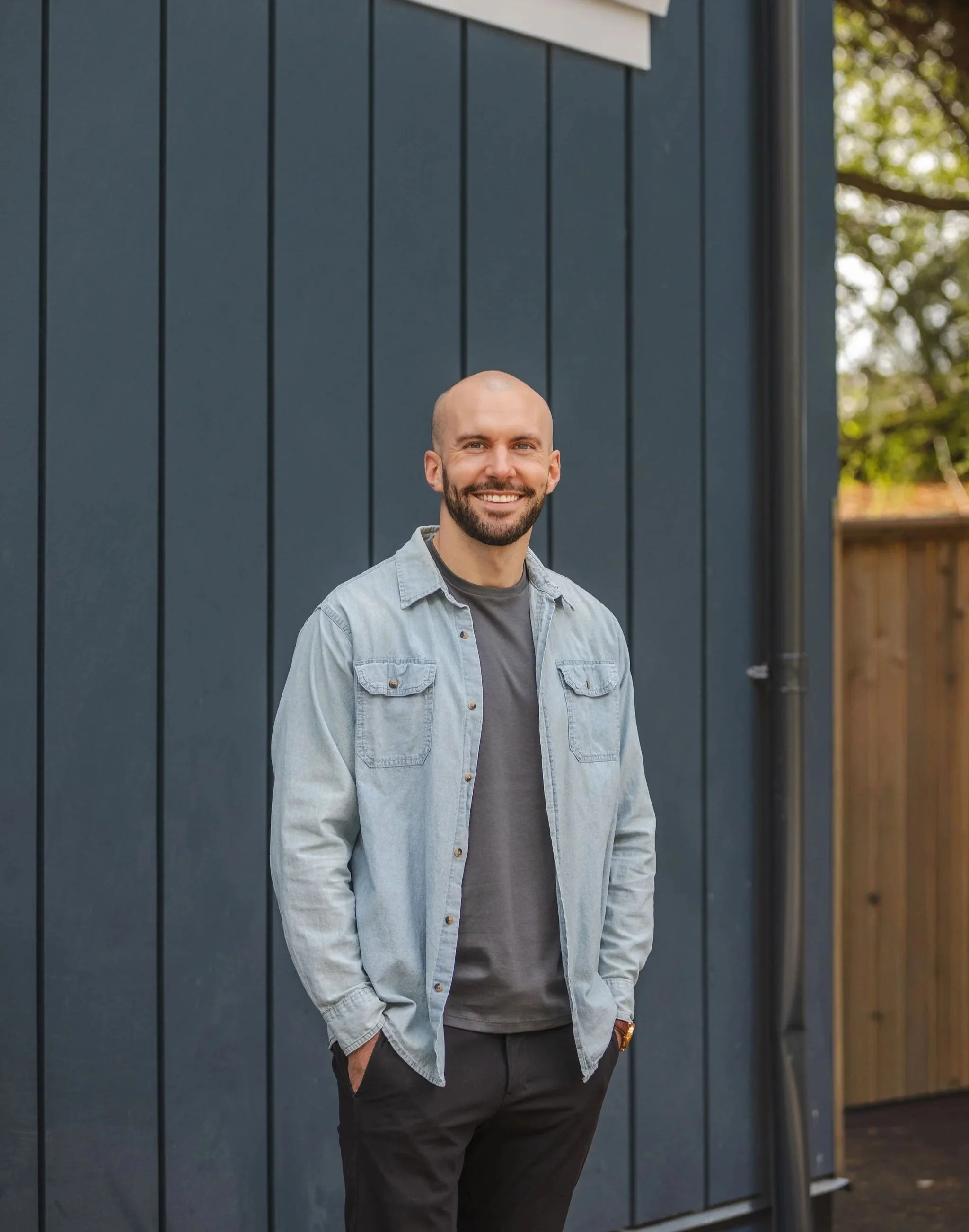 A smiling man with a beard and a shaved head, wearing a light denim jacket over a gray shirt, standing outdoors in front of a dark gray wooden wall with greenery and trees in the background.