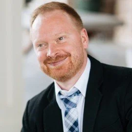 A man with red hair and a beard smiling in a professional setting, wearing a dark blazer and a light blue tie.