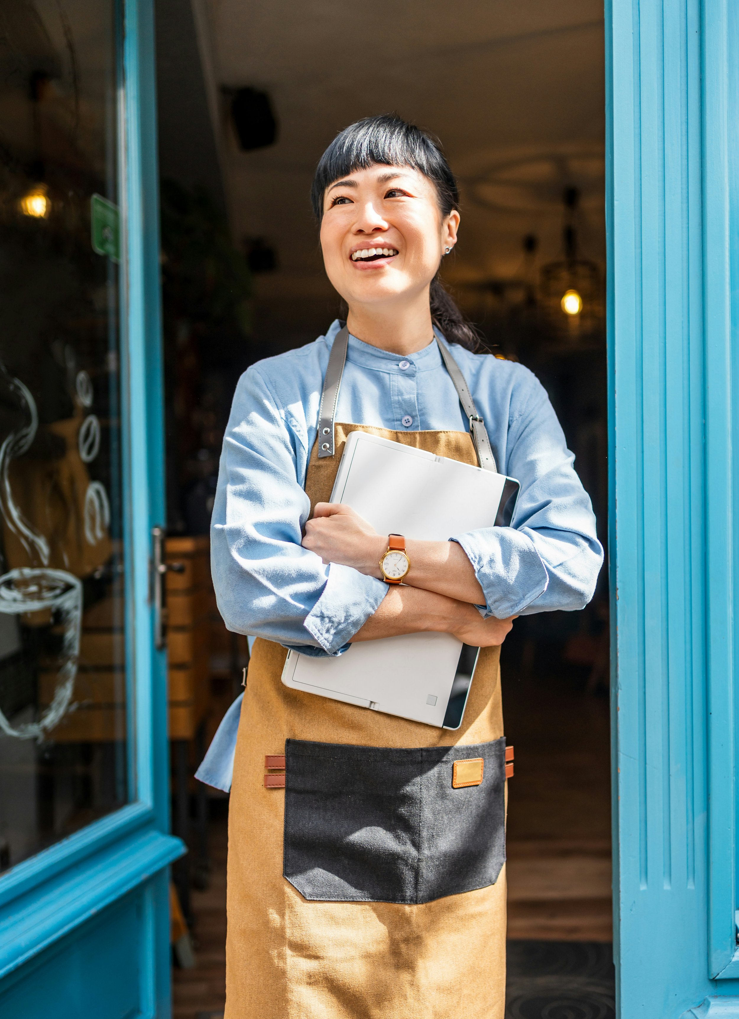 A woman standing at a doorway, smiling, holding a clipboard, wearing a light blue shirt, brown apron, and a watch. The door frame is painted blue.