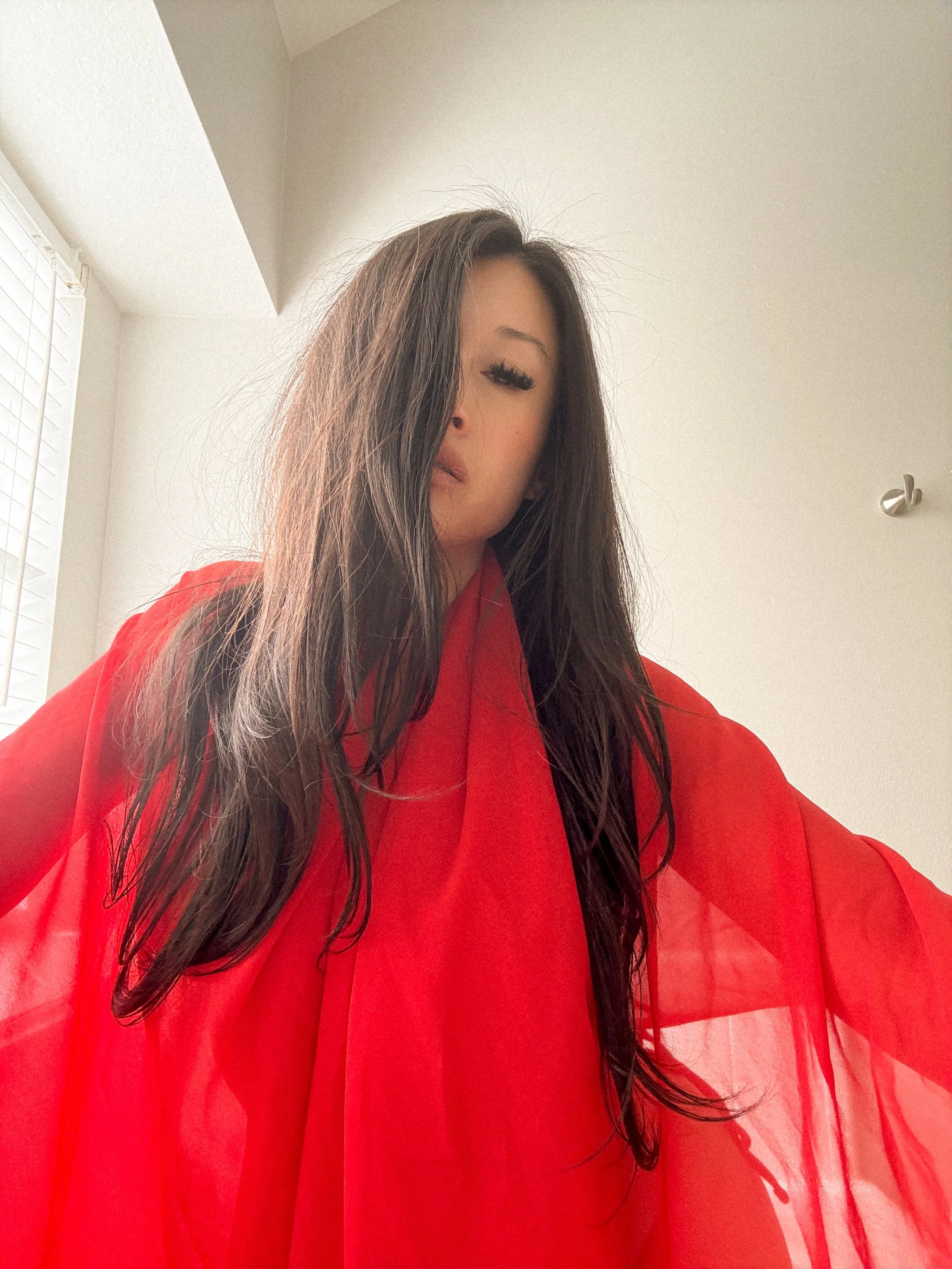 A woman with long dark hair, partially covering her face, wearing a red sheer garment, standing indoors with white walls and a window with blinds.