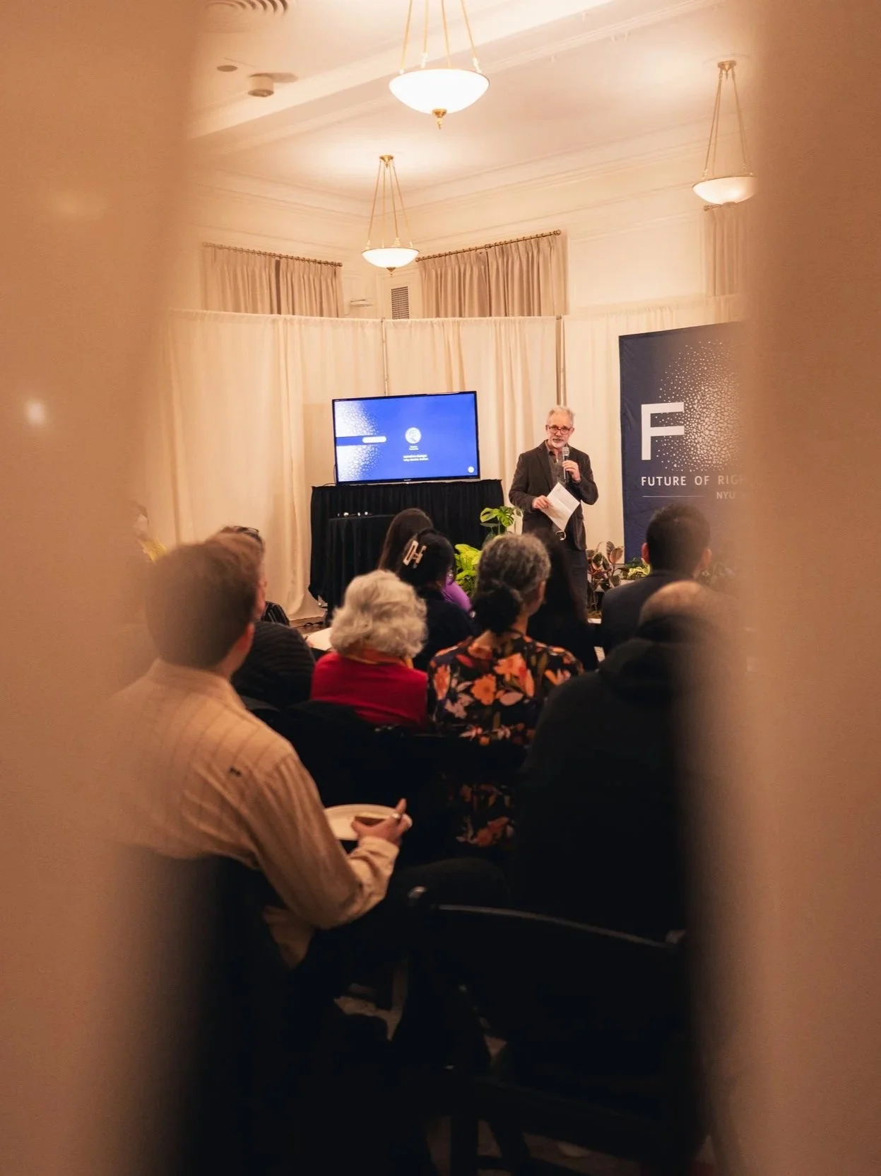 A speaker giving a presentation at a conference on the future of research at NYU, with an audience seated in a room with cream-colored curtains and chandeliers, viewed through a partially open doorway.
