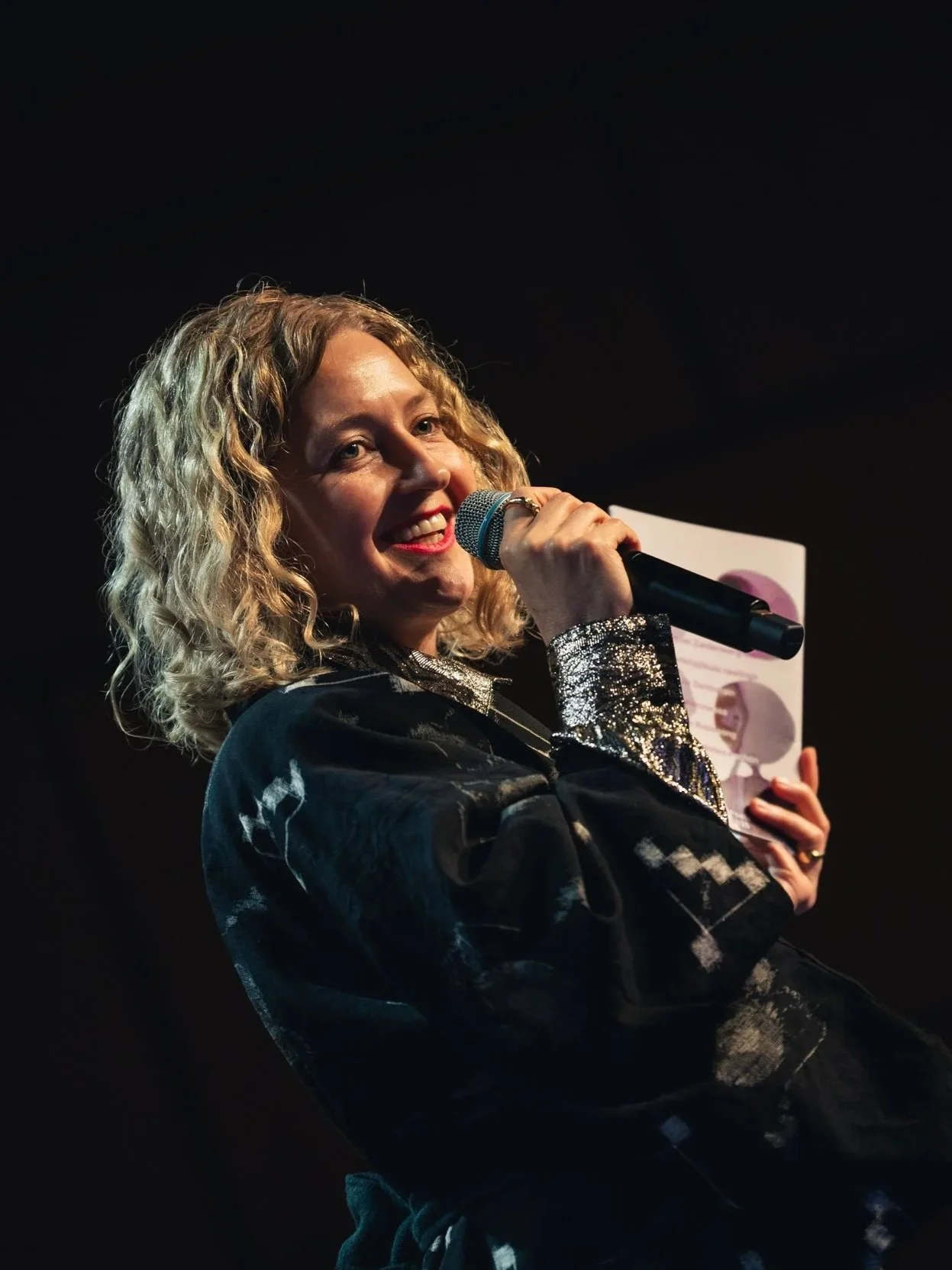 Woman with curly blonde hair holding a microphone and a paper, smiling on stage with dark background.