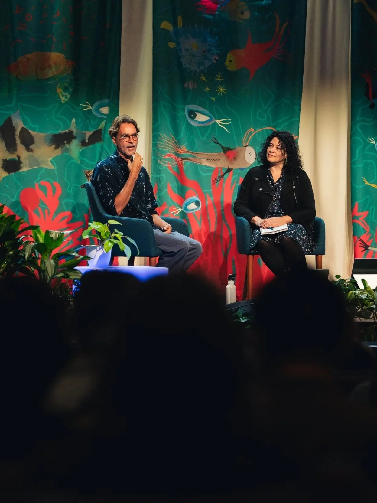 A man and a woman are sitting on a stage with colorful marine-themed backdrops featuring fish, coral, and other sea creatures. The man is speaking and gesturing with his right hand, while the woman listens with a notebook in her lap.