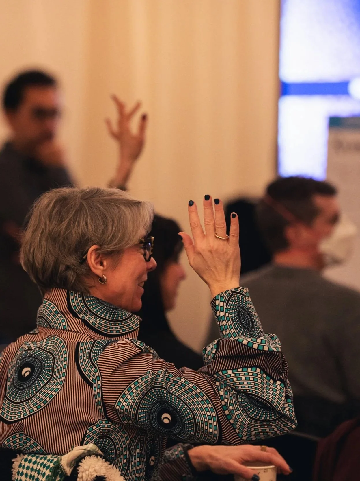 A woman with gray hair, glasses, and a patterned shirt raising her hand in a meeting or conference. Several other people are seated nearby, with some also raising their hands.