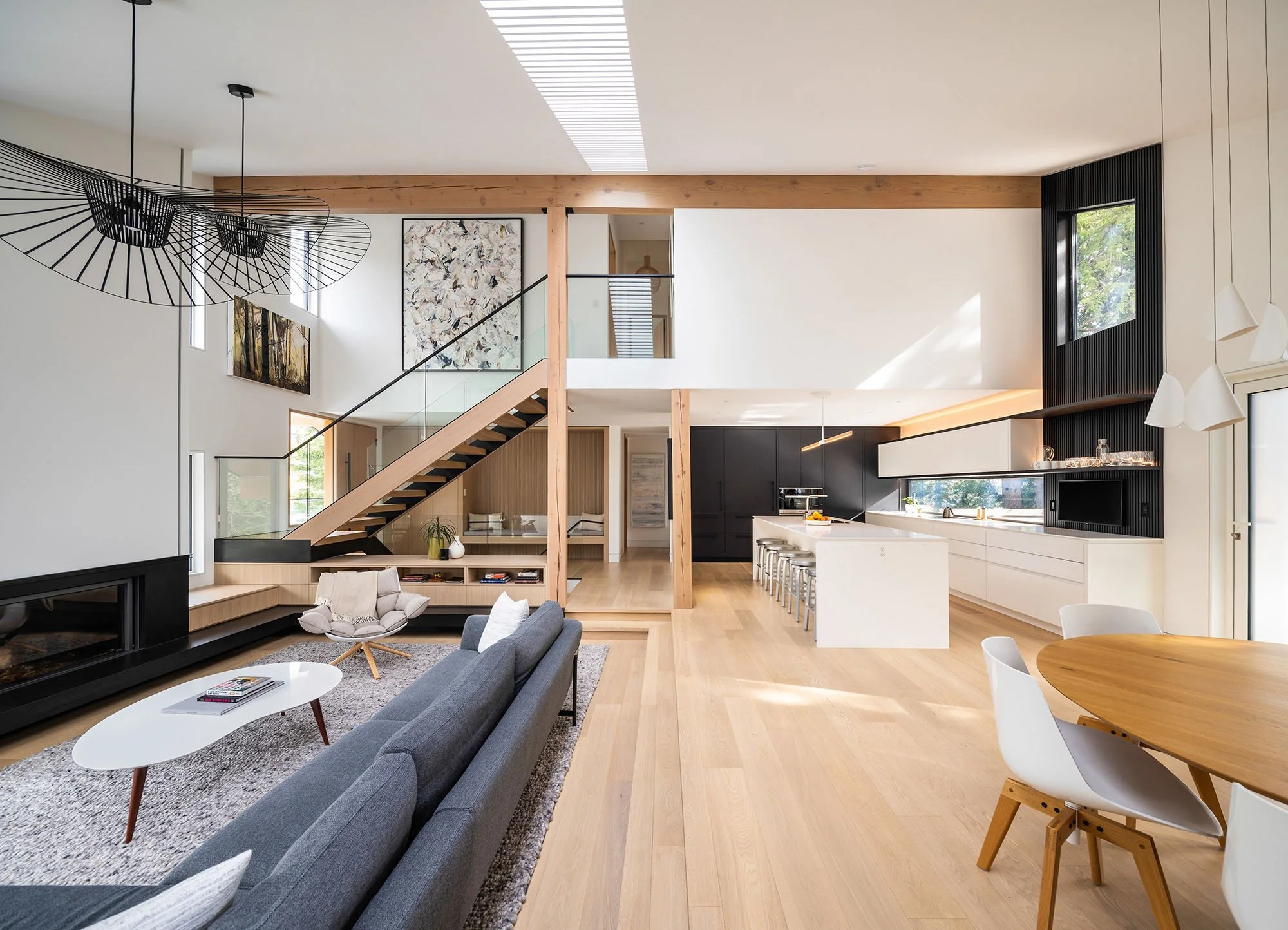 Open concept living room and kitchen with natural wood floors, modern furniture, black and white cabinetry, and large windows letting in natural light.