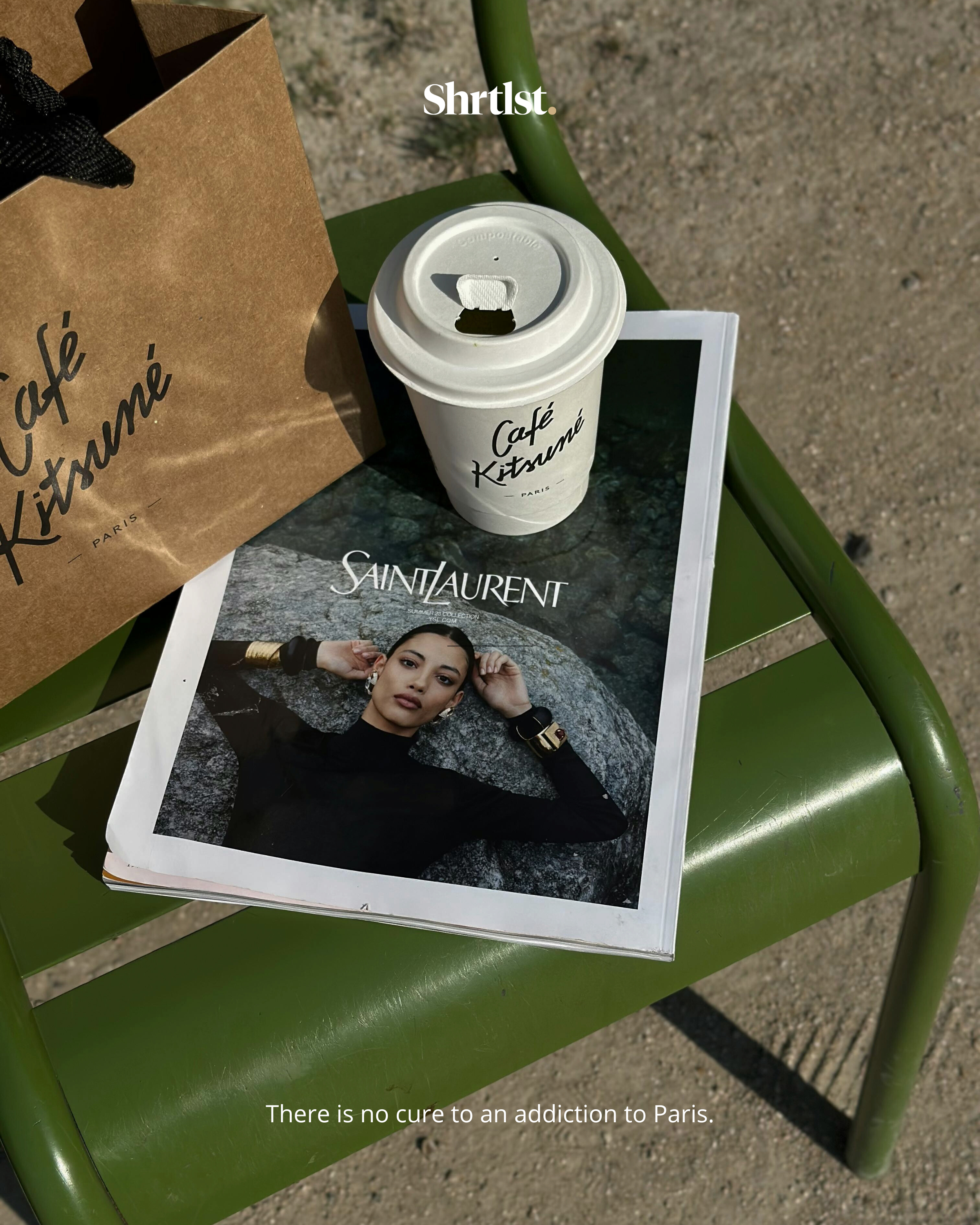 A green outdoor chair holding a Saint Laurent magazine, a takeout coffee cup with Café Kitsuné branding, and a brown paper bag with Café Kitsuné written on it. The ground is sandy. Text overlay reads, 'There is no cure to an addiction to Paris.'