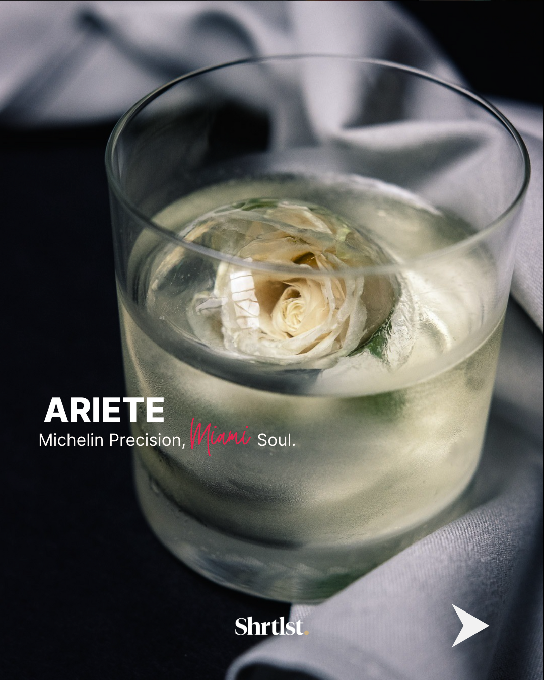 A glass of water with a white rose submerged in it, placed on a dark surface with gray cloth nearby.