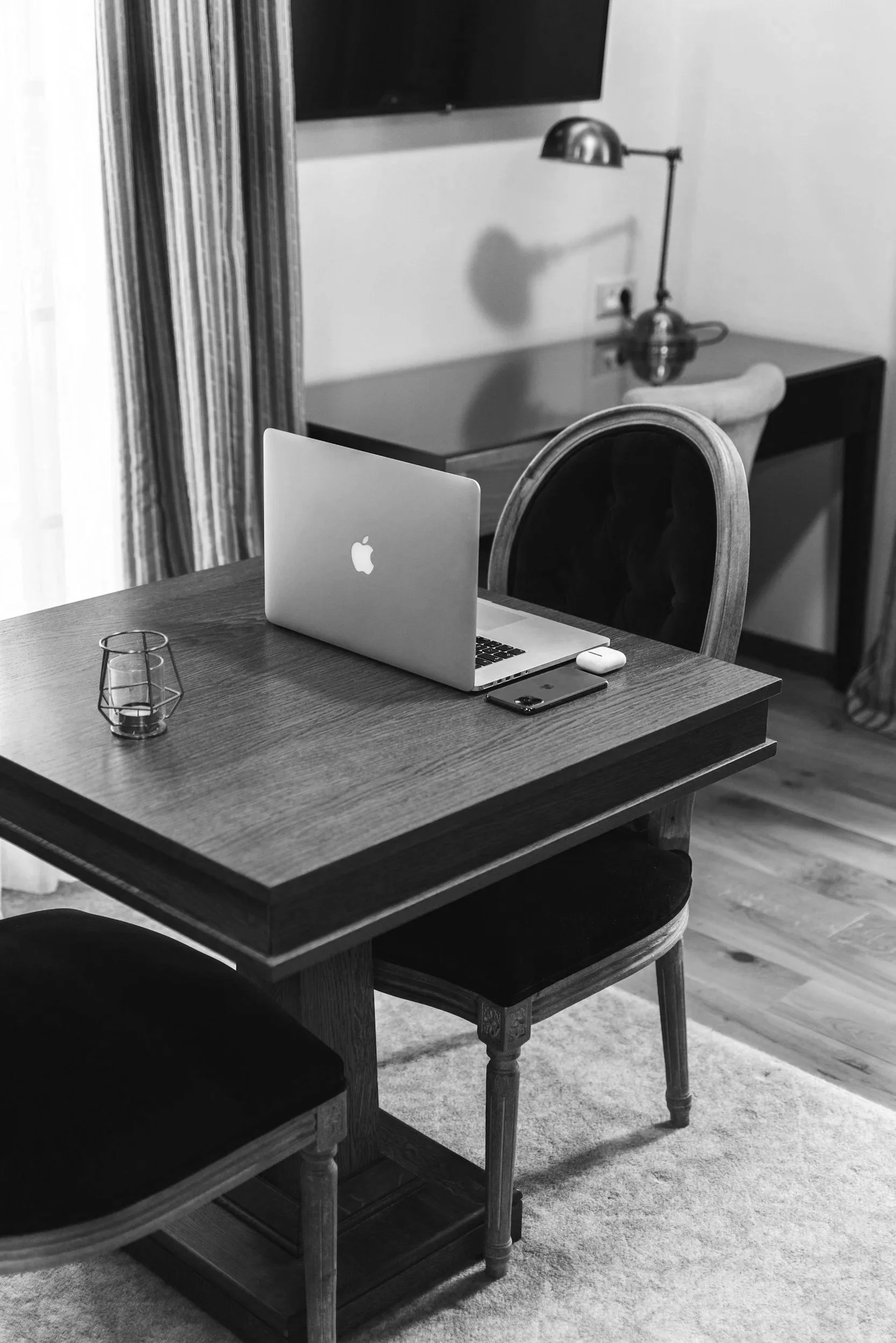 A black and white photograph of a dining room with a wooden table, chairs, and a laptop on the table. There is a lamp in the background, and a flat-screen TV on the wall.