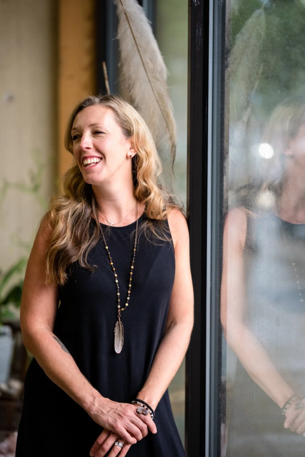 Smiling woman with long hair wearing a black dress and standing near a window, adorned with a feather necklace and bracelets.