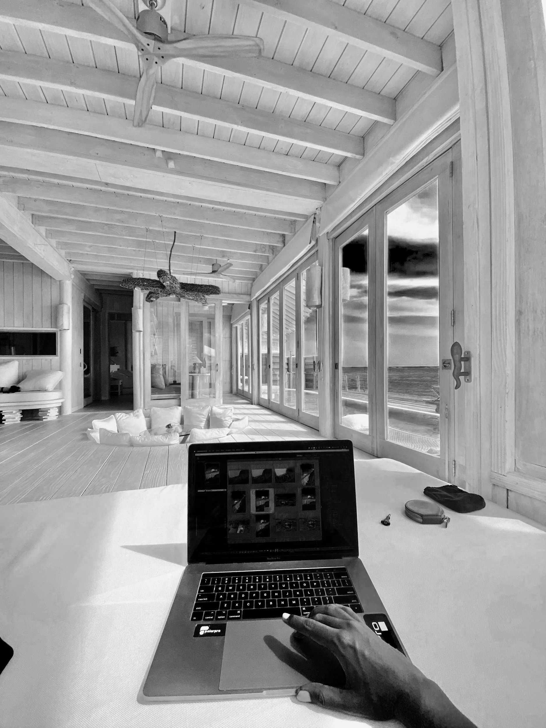 A black-and-white photo of a spacious room with large glass sliding doors revealing an ocean view. The room has a wooden ceiling with exposed beams and a ceiling fan. There is a laptop on a table in the foreground, and a hand resting on the laptop's trackpad. In the background, there are white cushions and a low table, with a glimpse of a bed in the left corner.
