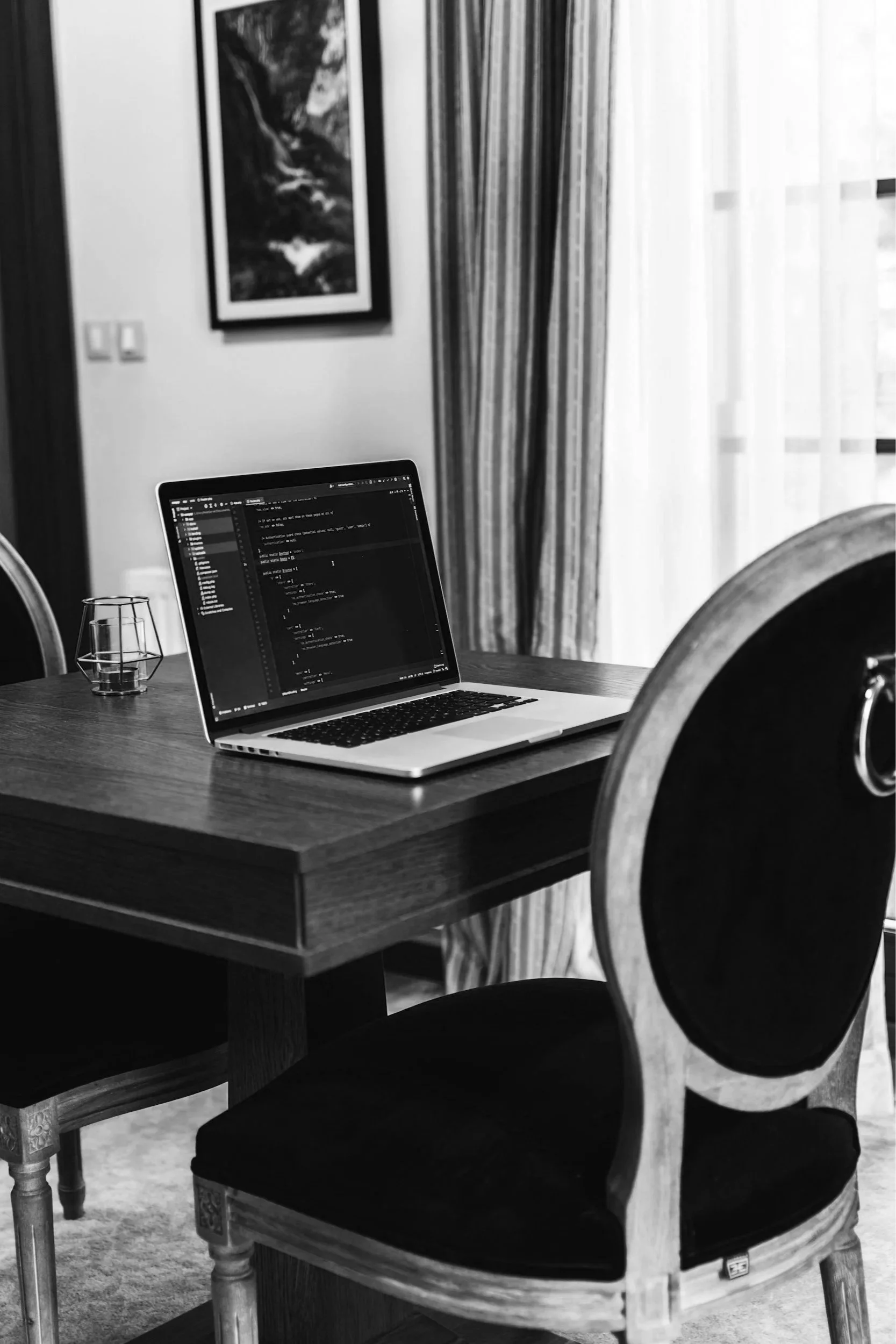 A wooden dining table with a laptop displaying computer code on its screen, a glass, and a chair in a room with curtains and wall art.