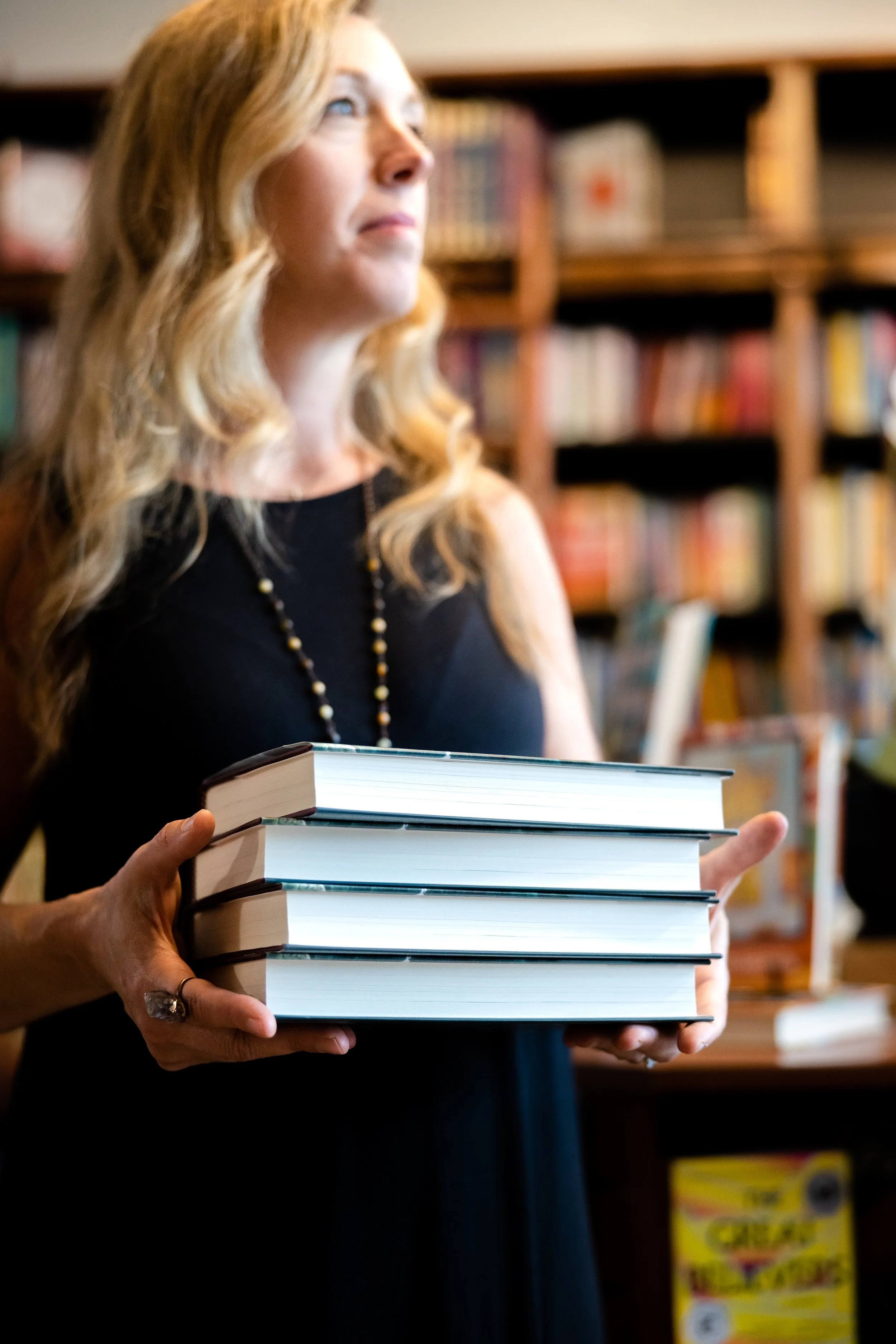A woman with long blonde hair holding a stack of books in a library.