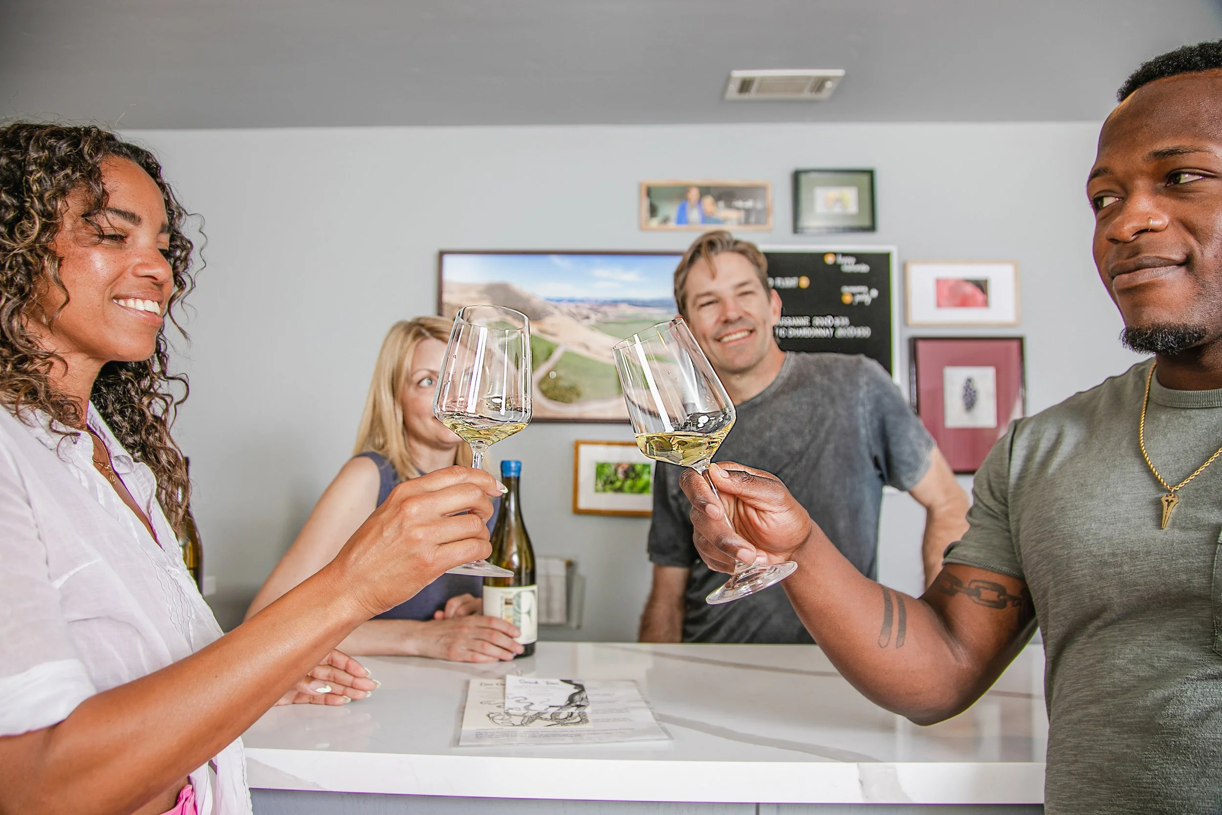 Group of four people in a wine tasting room clinking white wine glasses at a counter, smiling, with wine bottles and framed pictures in the background.