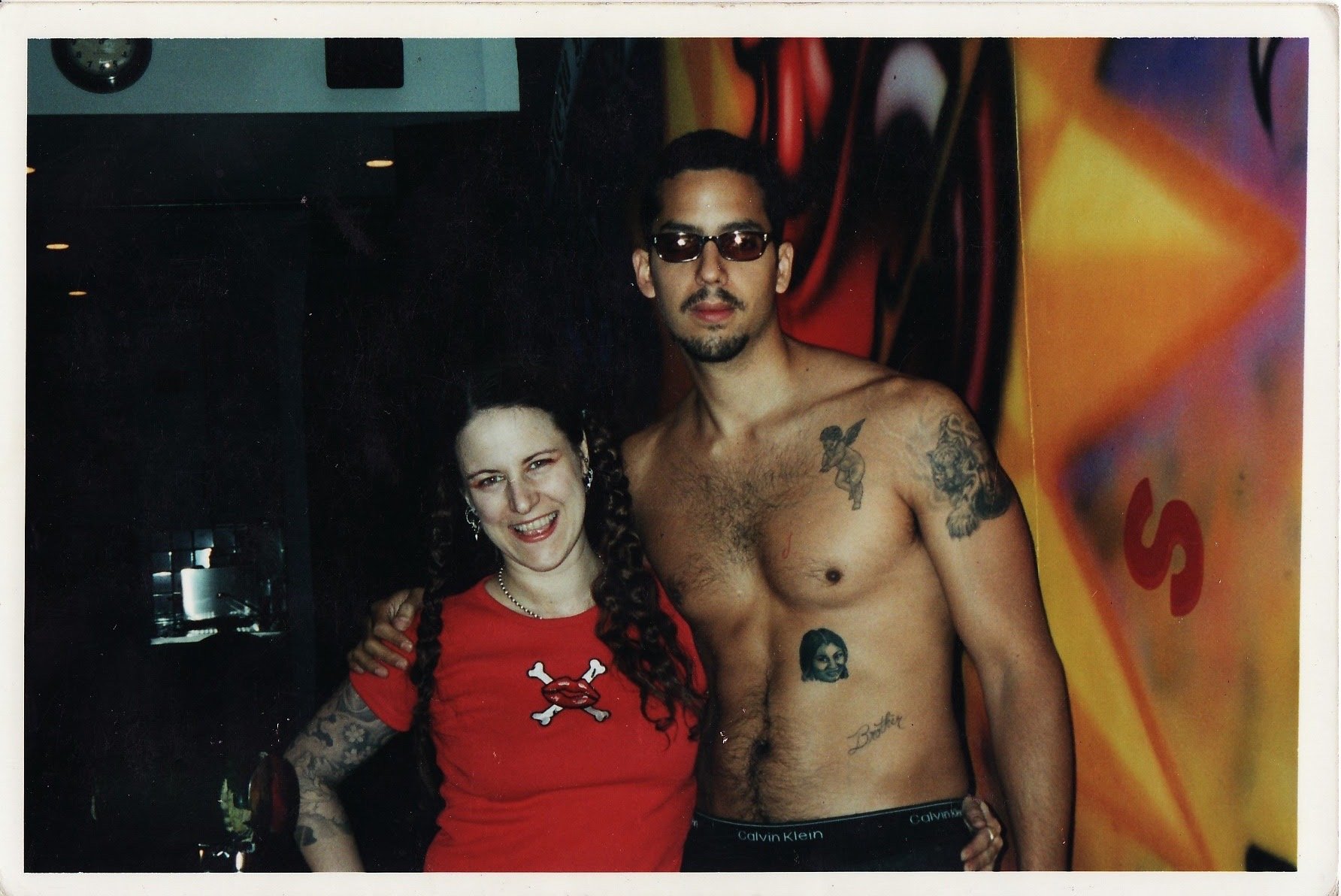 A woman, Ronda in a red shirt with a skull graphic stands next to a shirtless man with tattoo, David Blaine Background includes colorful graffiti.