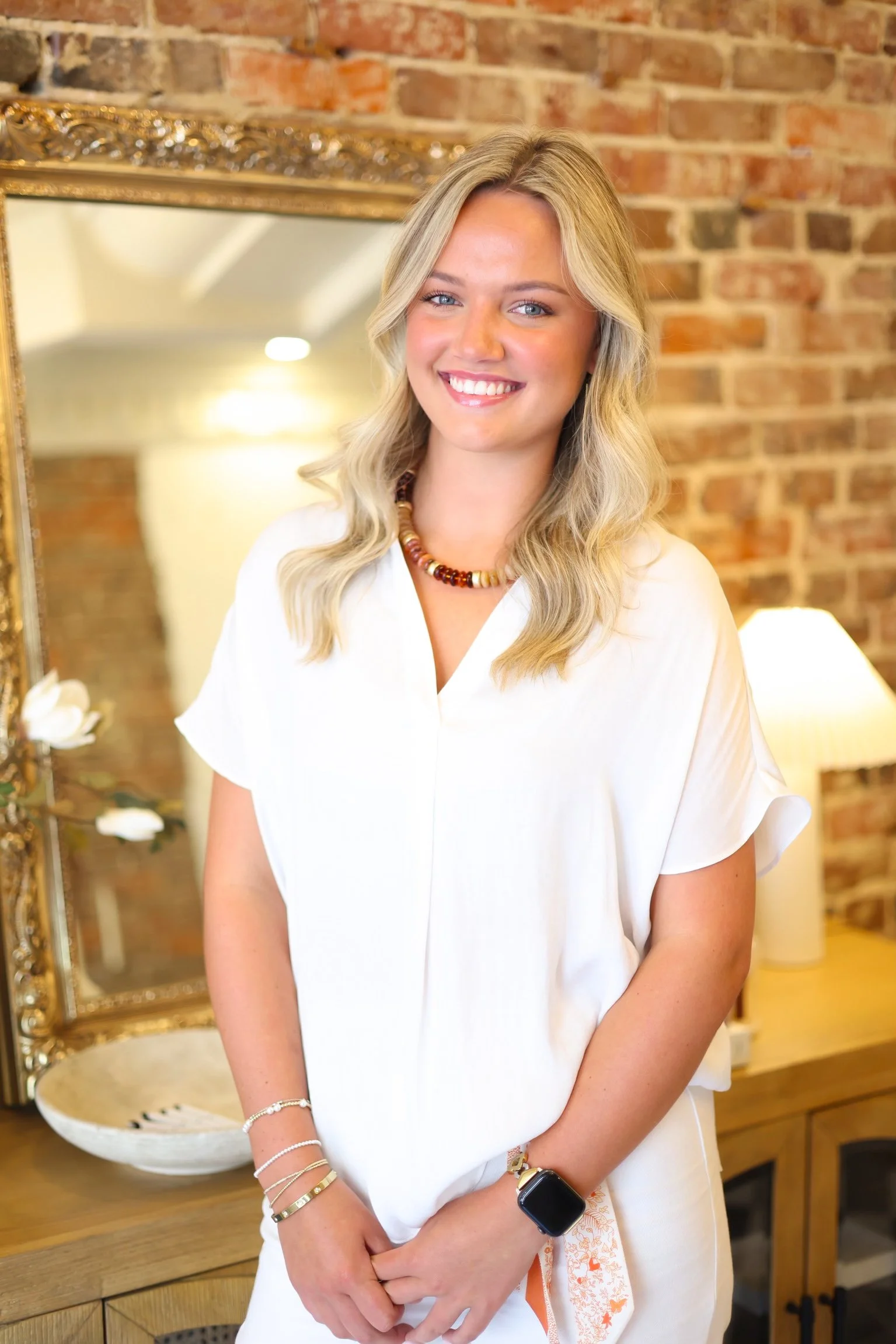 A woman with long blonde hair, blue eyes, and a bright smile sitting on a dark green sofa against a brick wall. She is wearing a dark sleeveless top, white pants, a pearl ring, and multiple bracelets, with her hand resting under her chin. There is a gold-colored side table next to her.