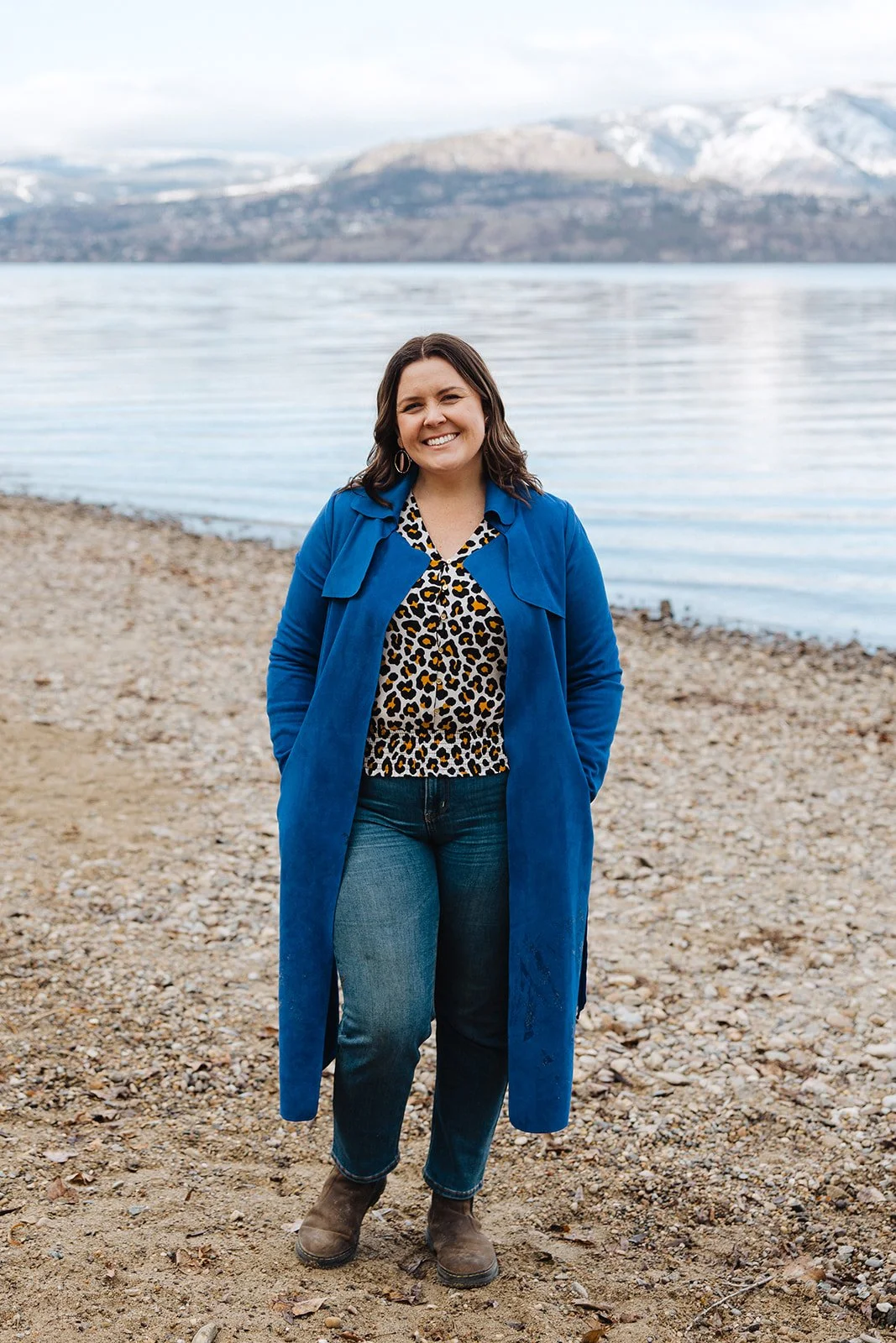 A woman standing on a rocky beach near a body of water, with mountains in the background, smiling and wearing a blue coat and leopard print top.